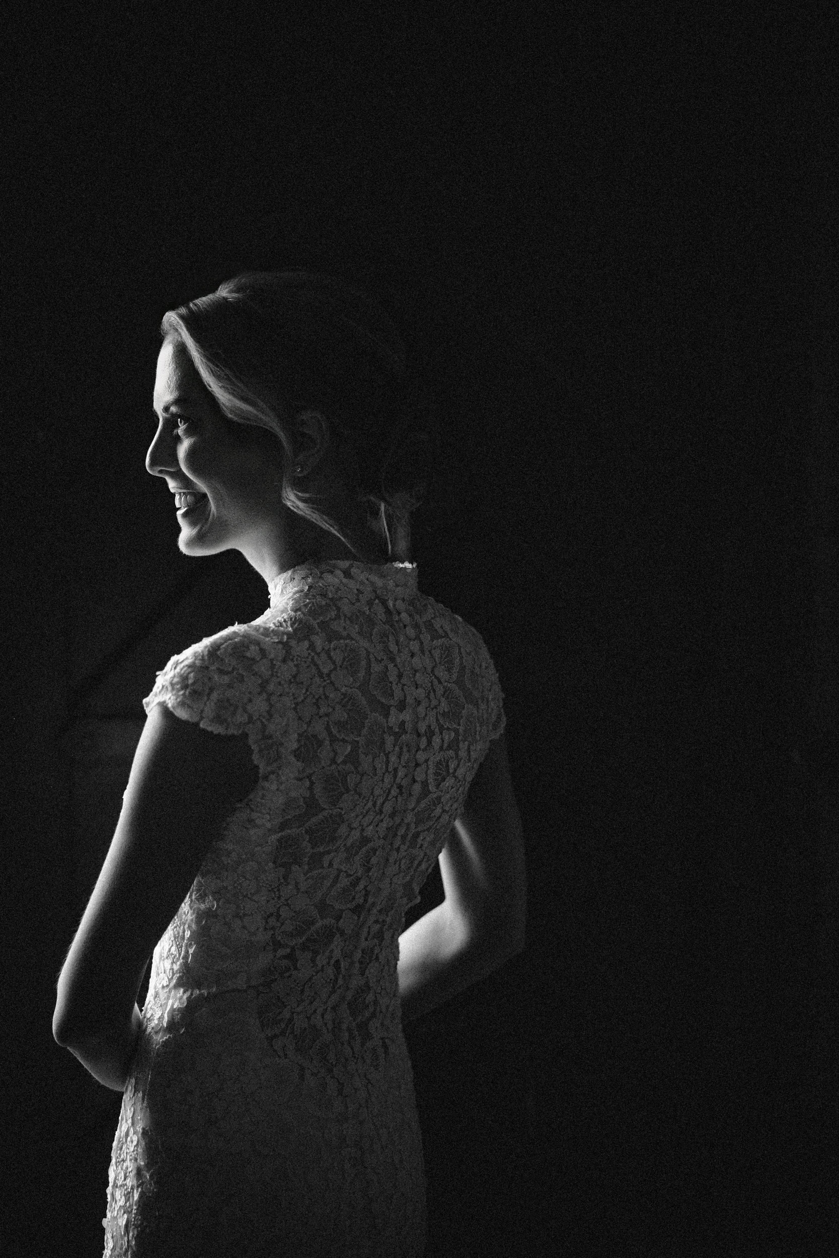 A bride in a lace dress stands in profile, partially lit against a dark background at a Houston Hall wedding, her hair styled up and a slight smile on her face.