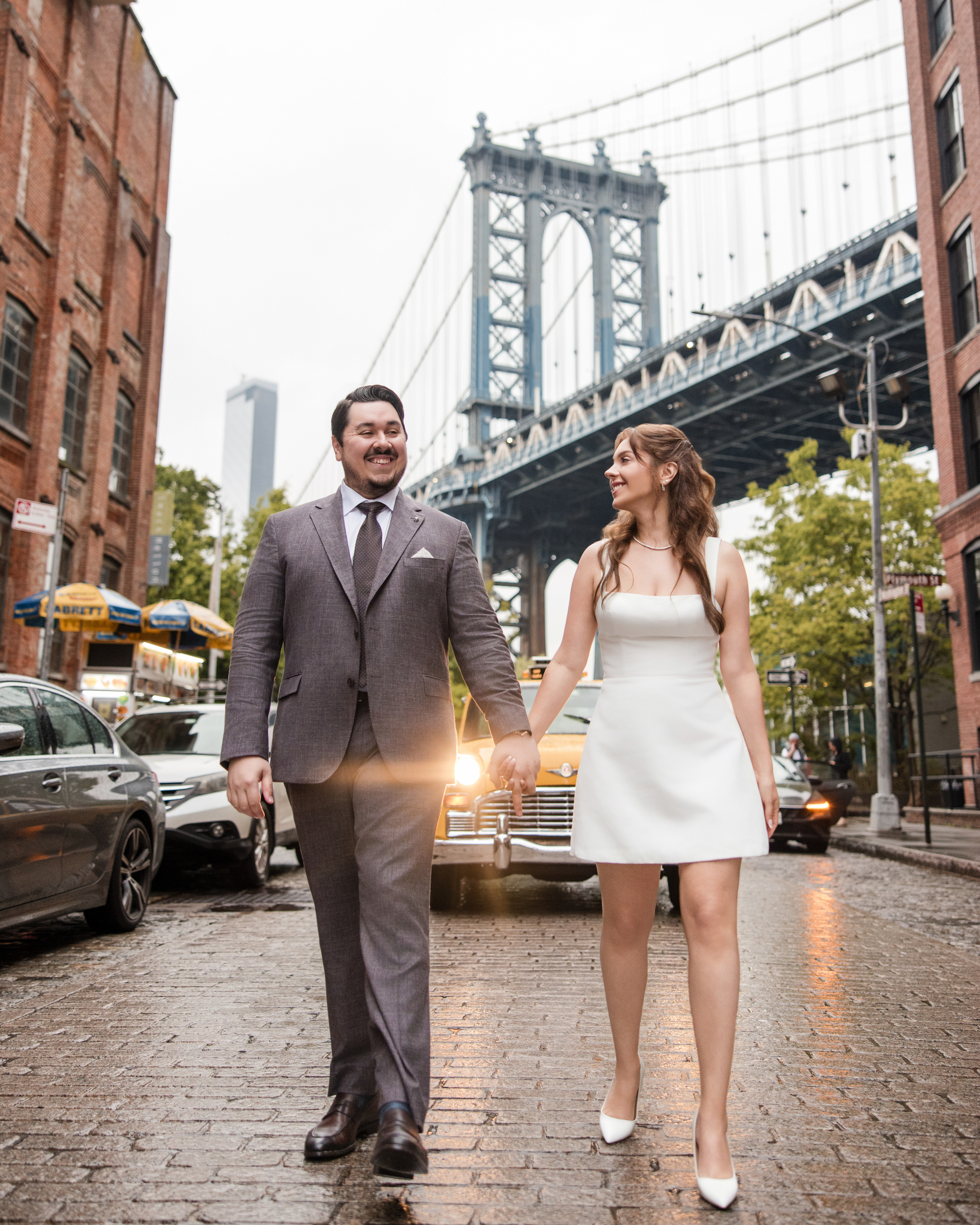 A couple dressed formally walks hand in hand on a cobblestone street during a rainy NYC engagement session in front of the Manhattan Bridge.