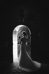A woman in a lace wedding dress stands in a doorway at Houston Hall, framed by light streaming in from outside, her back to the camera.
