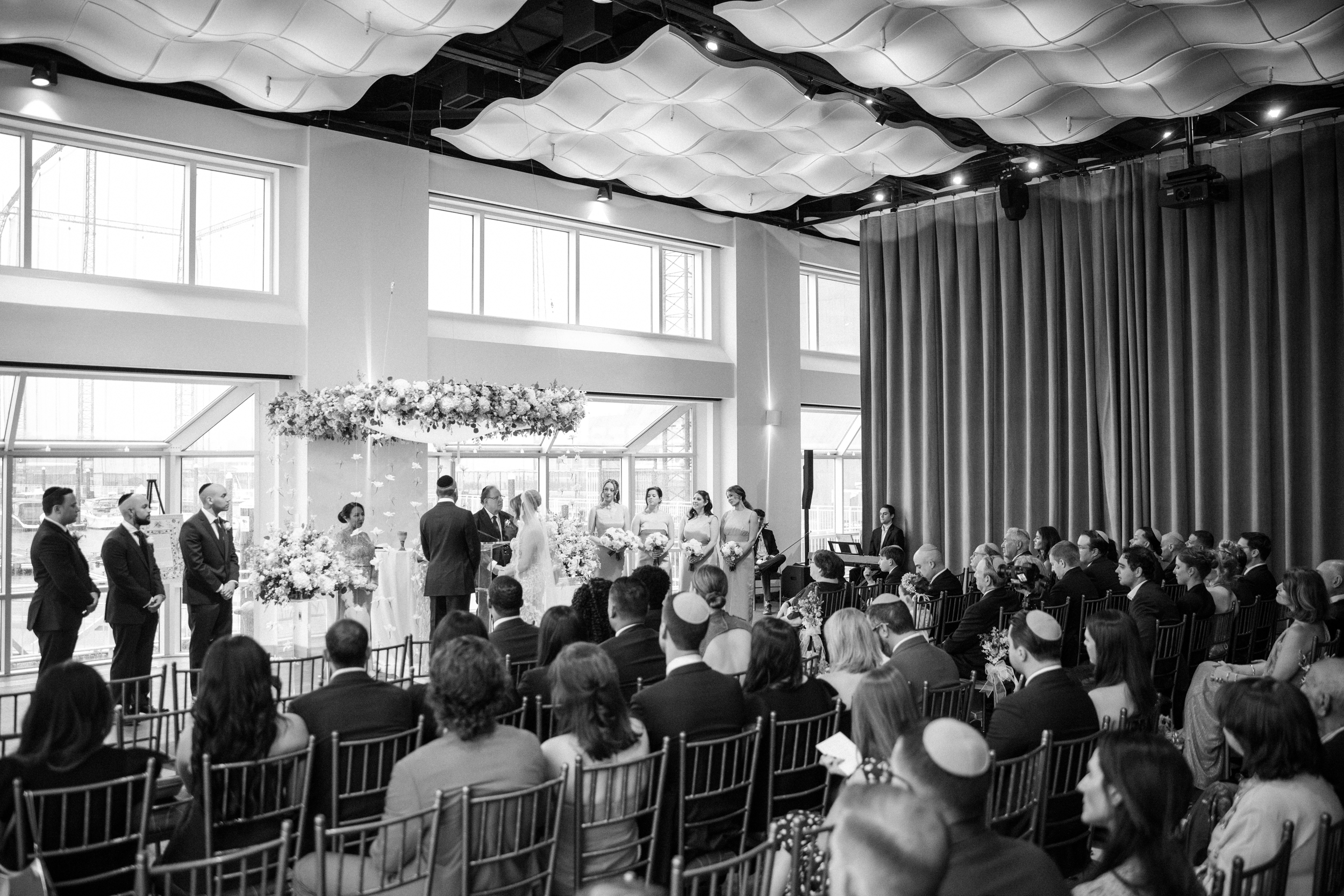 A wedding ceremony in progress indoors at Pier Sixty and Current, with a couple standing under a floral canopy, an officiant, wedding party members, and seated guests observing.