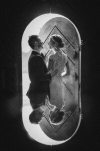 A couple in formal attire stands facing each other in front of an arched doorway at their Houston Hall wedding, their reflection visible on a shiny surface below.