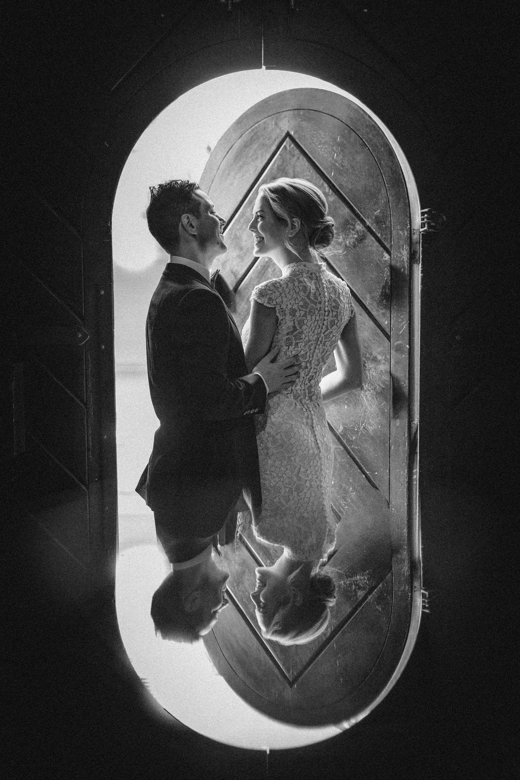 A couple in formal attire stands facing each other in front of an arched doorway at their Houston Hall wedding, their reflection visible on a shiny surface below.