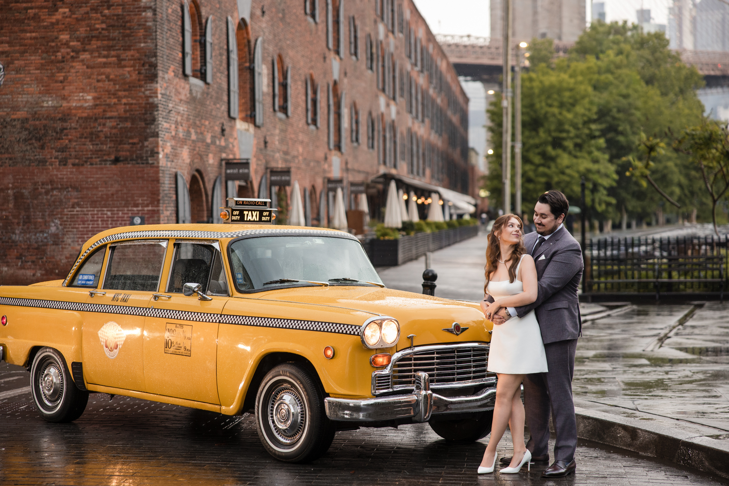 A couple stands close together on a wet street beside a vintage yellow taxi, with brick buildings and a bridge in the background, perfectly capturing a romantic rainy NYC engagement session. A couple stands close together on a wet street beside a vintage yellow taxi, with brick buildings and a bridge in the background, perfectly capturing a romantic rainy NYC engagement session.