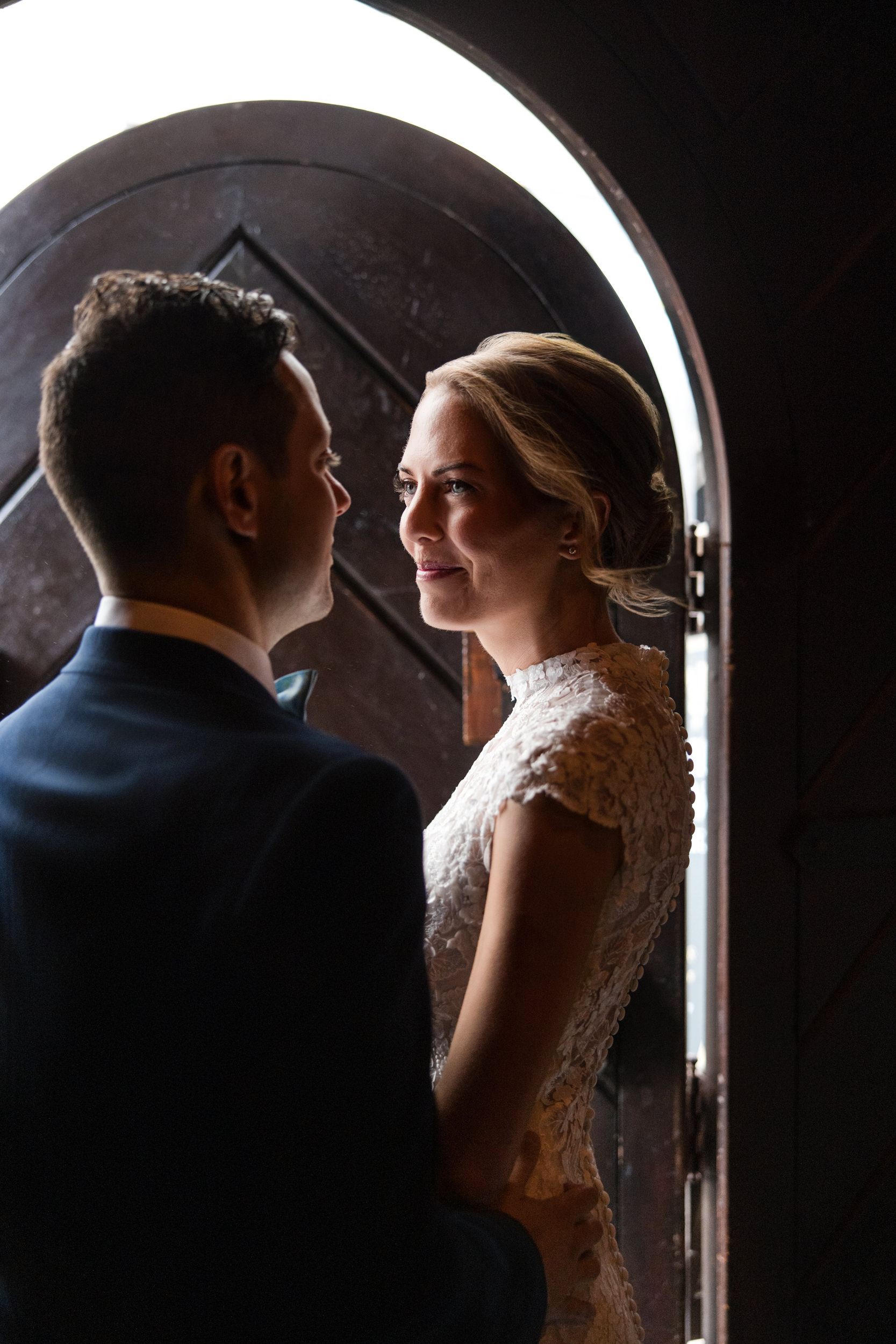 A bride and groom stand facing each other near a wooden arched doorway at their Houston Hall wedding, with soft light illuminating their faces.