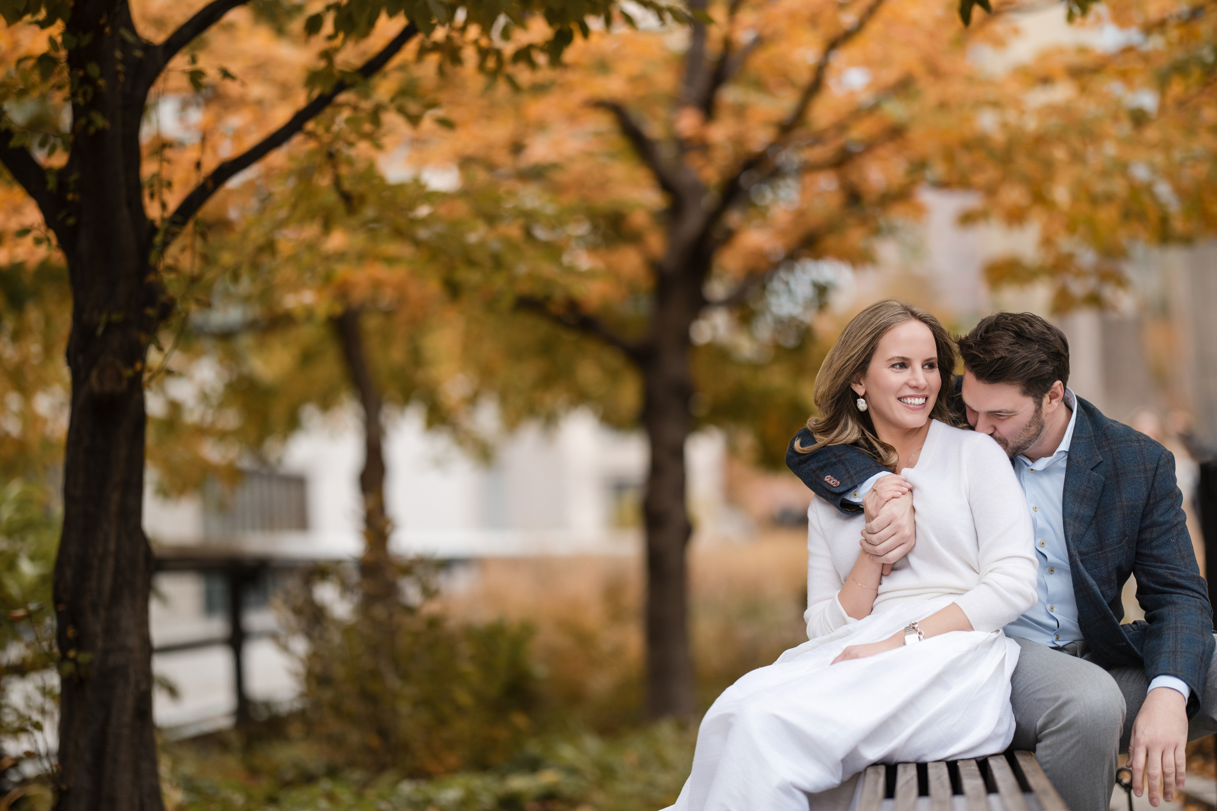 A couple sits closely together on a bench in a park with autumn foliage, smiling and holding hands during their High Line engagement session.