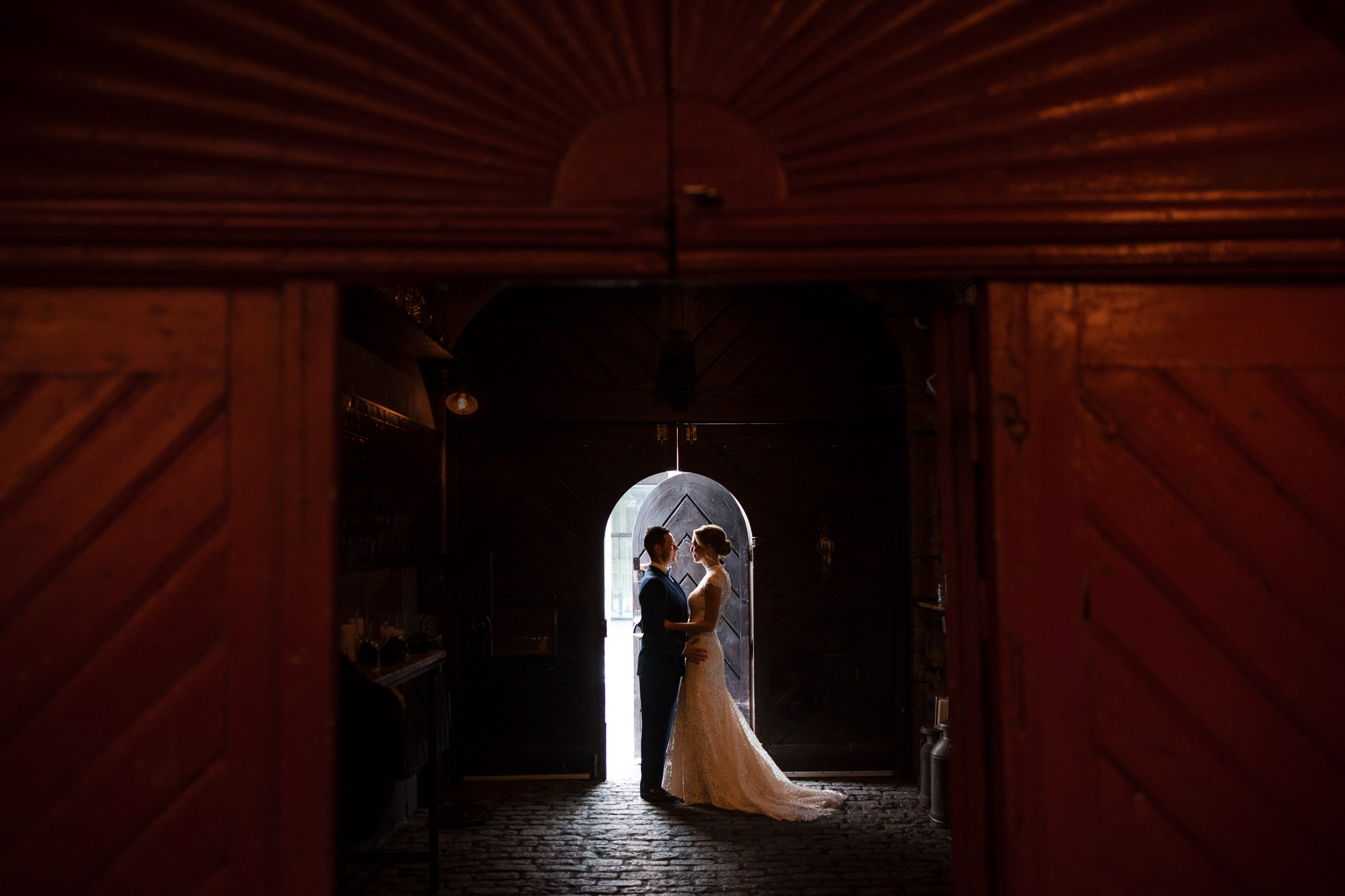 A bride and groom stand close together under an umbrella in a dimly lit, rustic doorway at their Houston Hall wedding, silhouetted by light from outside.