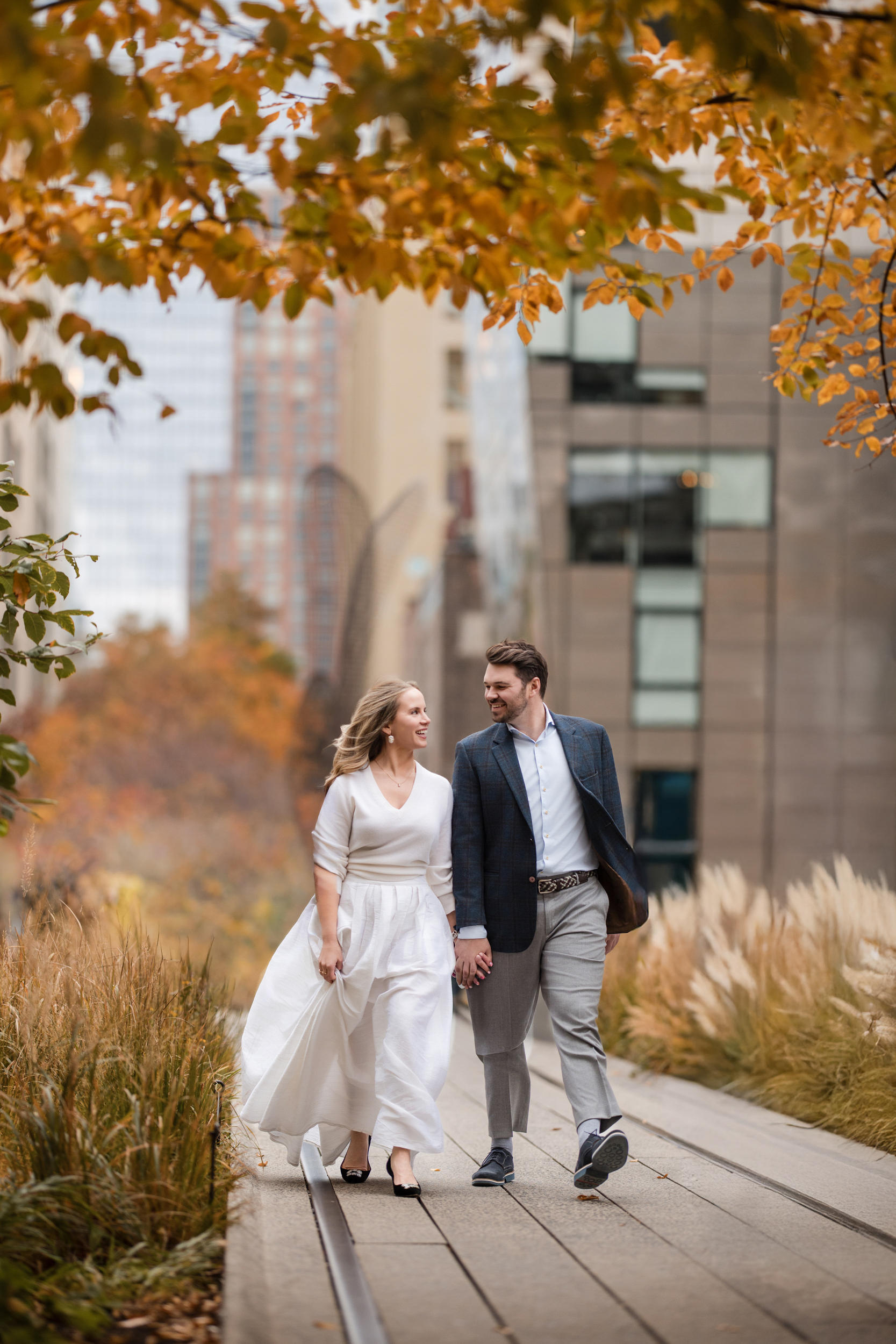 A couple walks hand in hand along the High Line during their engagement session, surrounded by autumn foliage and modern city buildings.