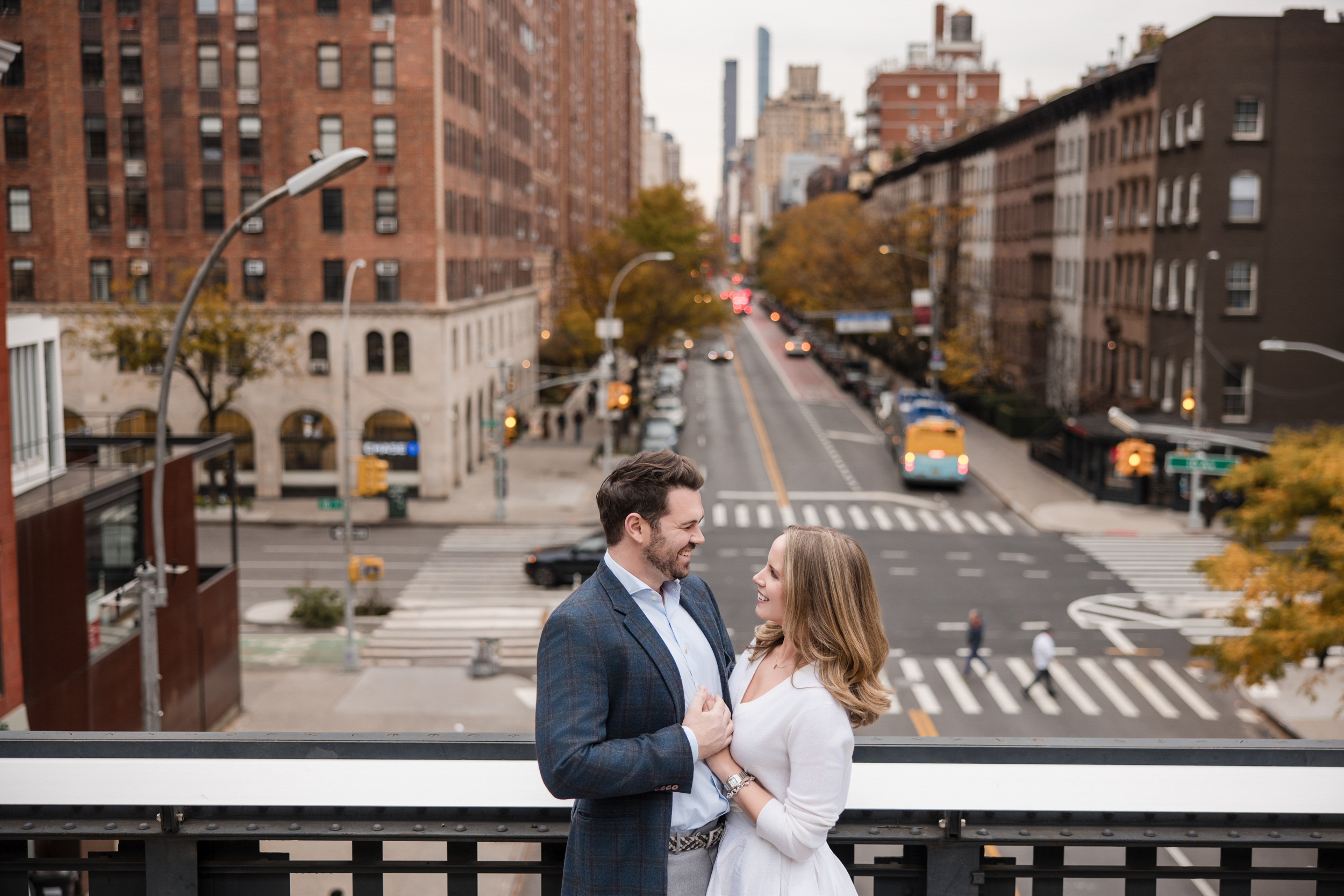 A man and a woman stand together on an elevated walkway overlooking a city street, capturing a special moment during their High Line engagement session with buildings, cars, and trees in the background.