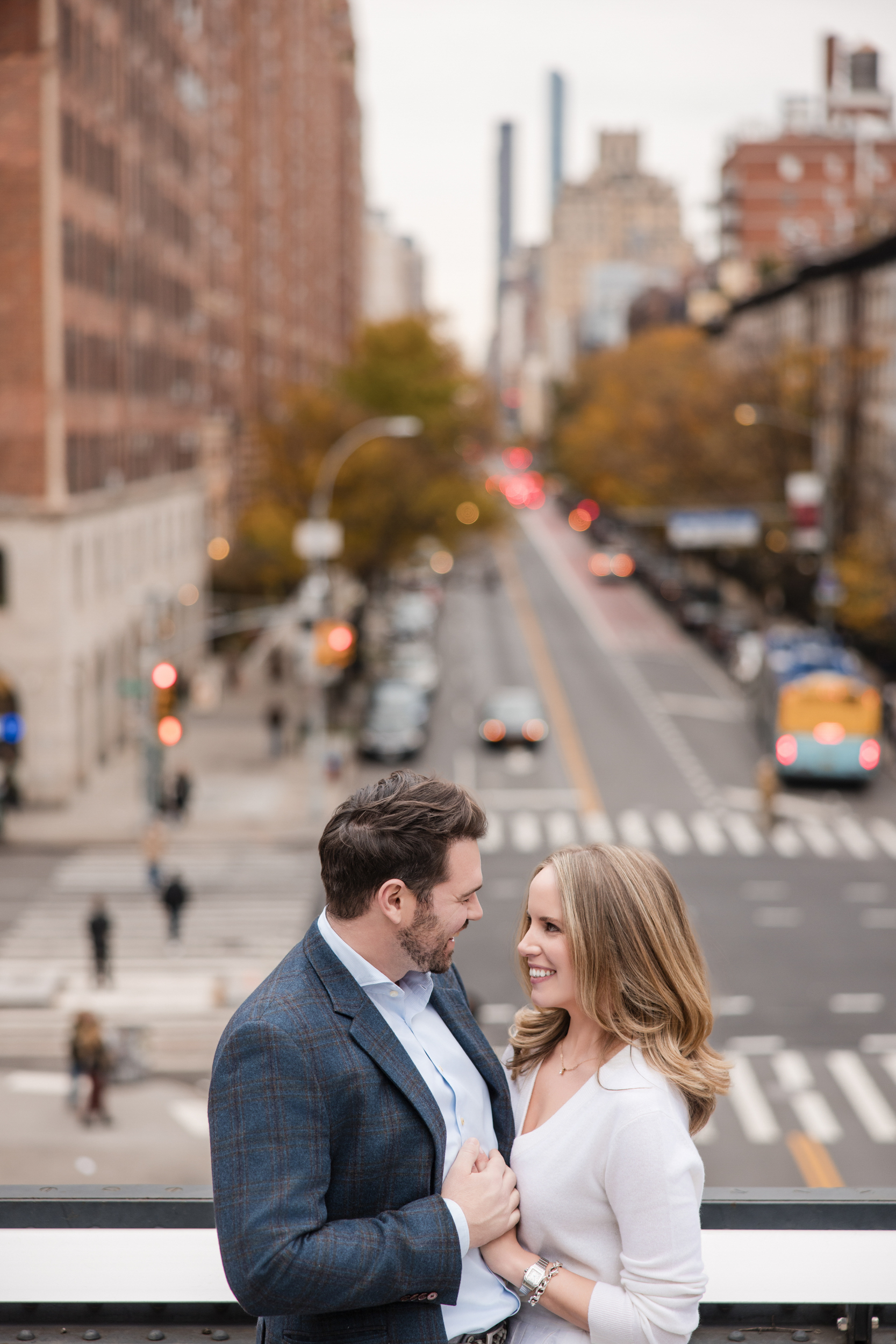 A man and woman stand close together on a city overpass during their High Line engagement session, looking at each other and smiling with a busy street and buildings in the background.