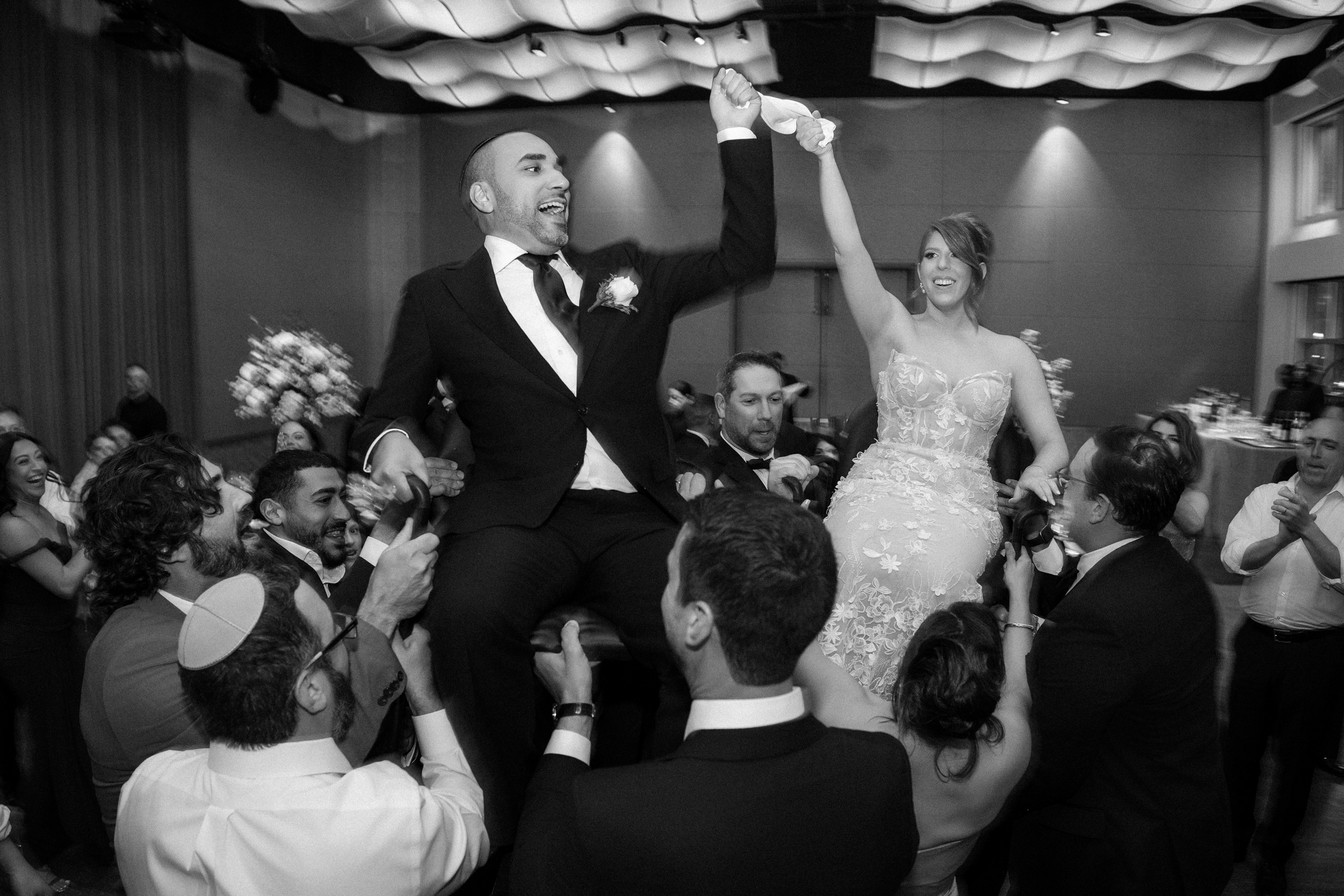A bride and groom are lifted on chairs by guests during a joy-filled Pier Sixty and Current Wedding, surrounded by cheering people in a grand banquet hall.