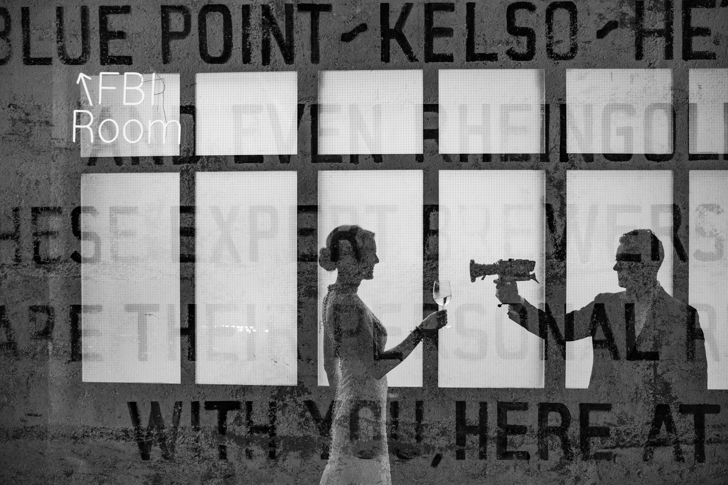Two people stand in front of large windows at a Houston Hall wedding; one holds a wine glass while the other playfully points a hair dryer at them. Overlay text includes “FBI Room” and other partially visible words.