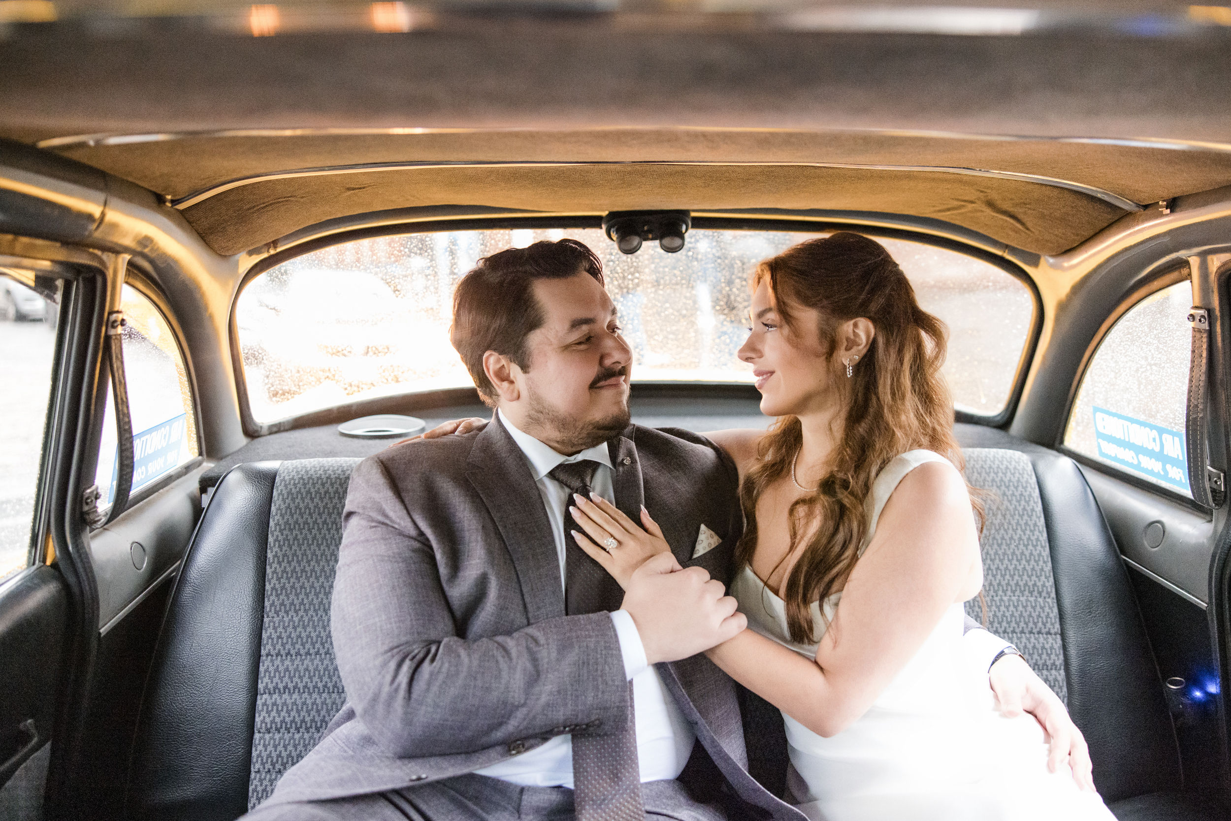 A couple in formal attire sits closely together in the backseat of a car, gazing at each other and holding hands during their rainy NYC engagement session.
