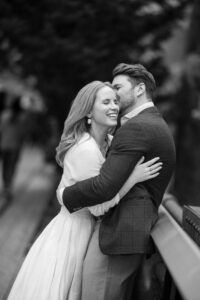 A couple embraces and smiles while standing by a railing outdoors during their High Line engagement session. The image is in black and white.