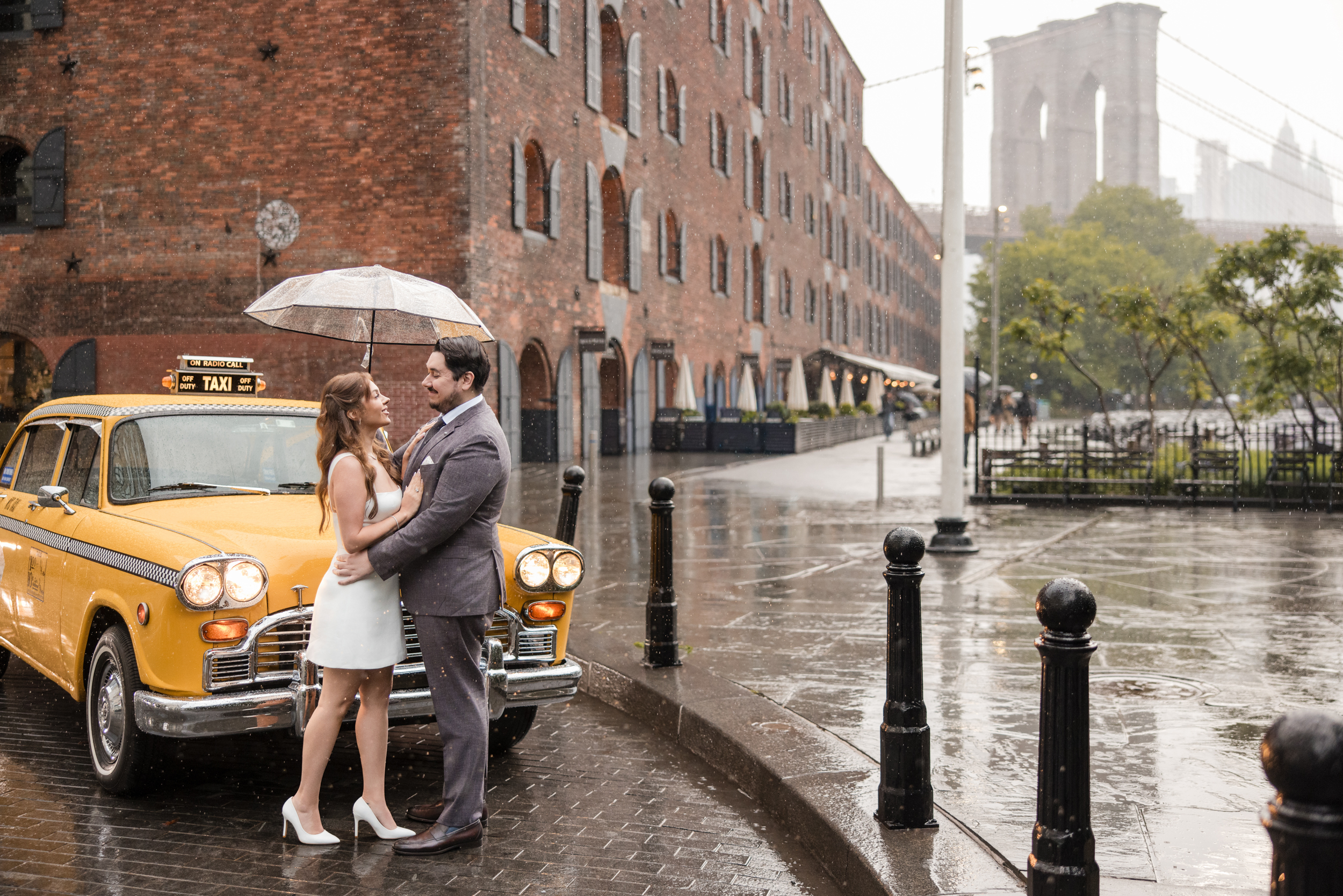 A couple stands under an umbrella in front of a vintage yellow taxi on a rainy street during their rainy NYC engagement session, with the Brooklyn Bridge visible in the background.