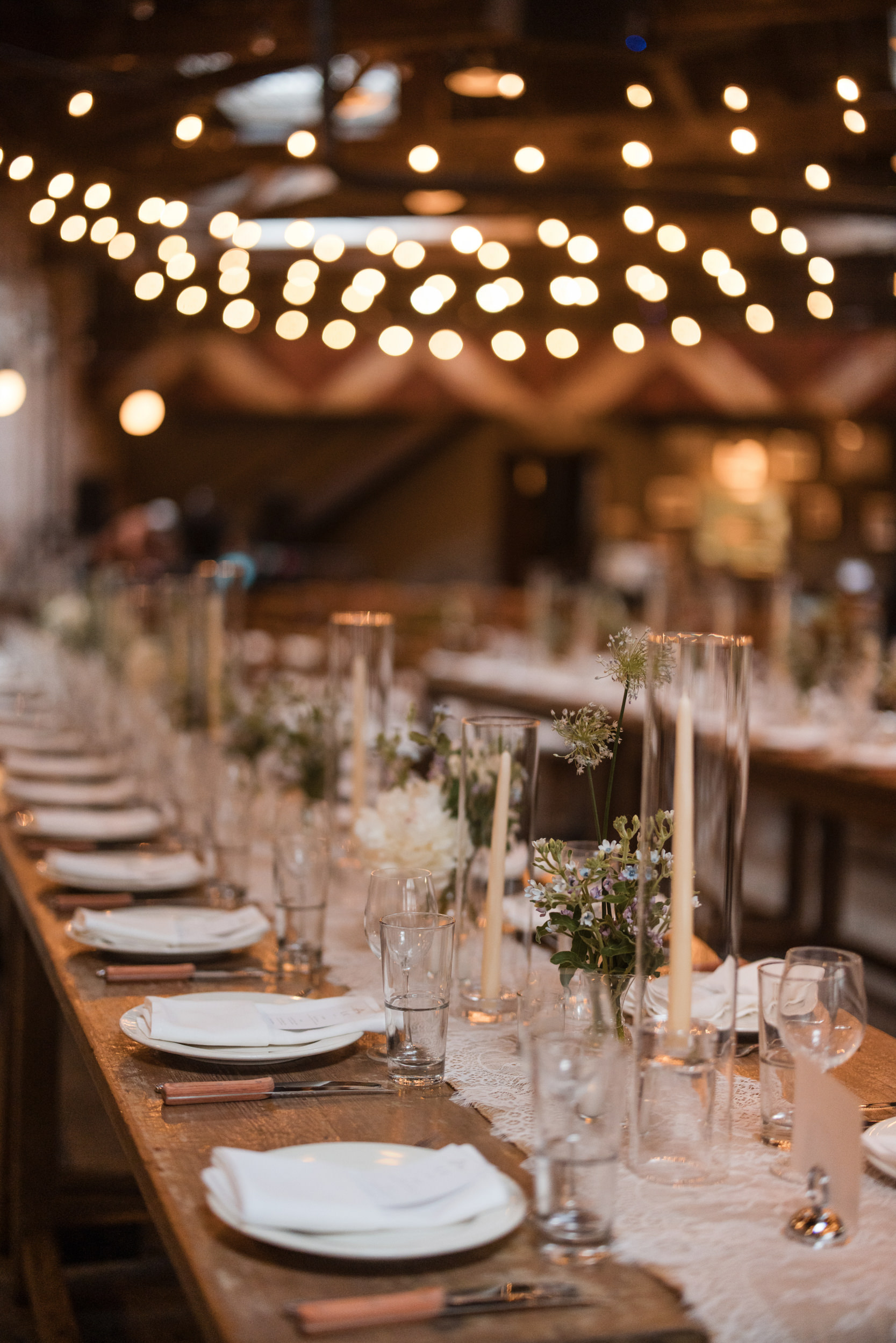 A long wooden table set with plates, glasses, cutlery, flowers, and candles in a warmly lit Houston Hall wedding venue with string lights overhead.