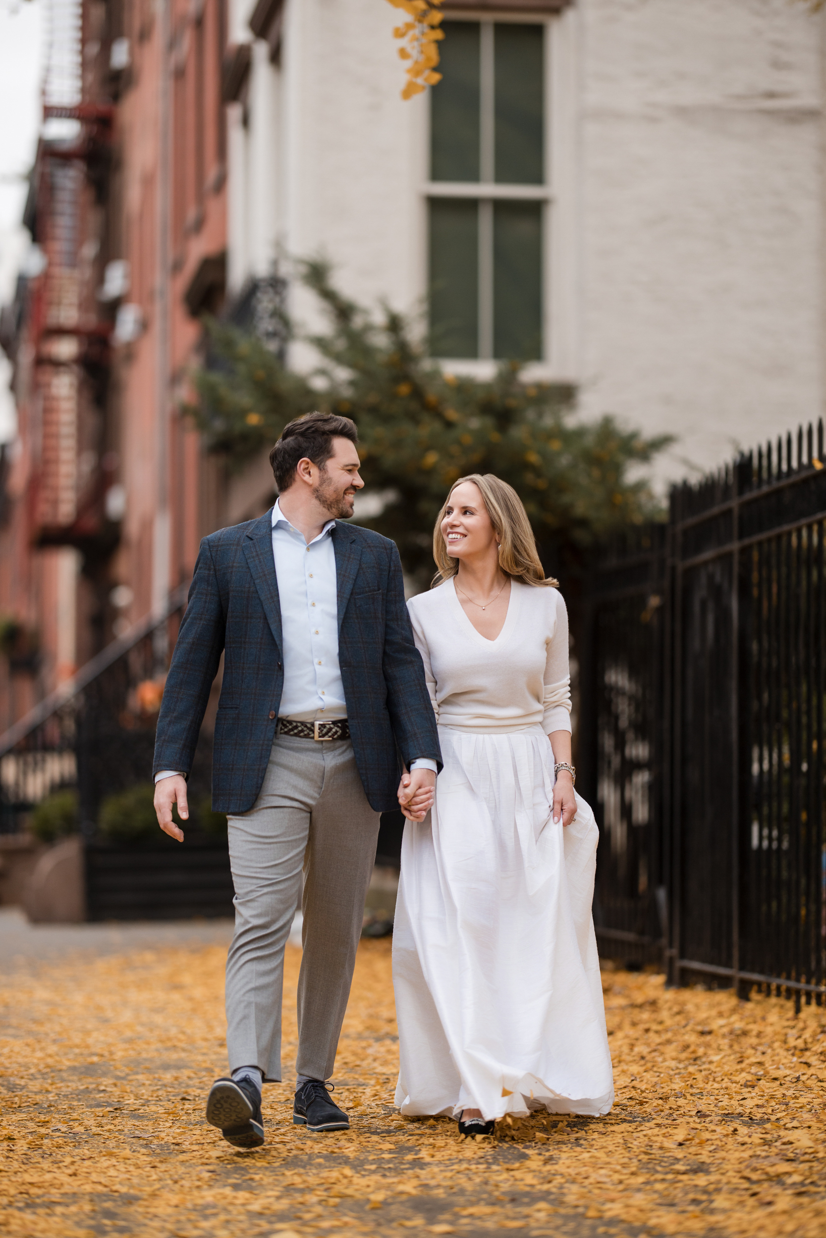A man and woman walk hand in hand on a city sidewalk covered in yellow leaves during a Chelsea engagement session, both dressed in semi-formal attire and smiling at each other.