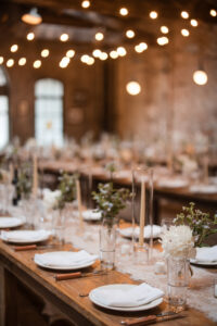 A long wooden table is set with white plates, glassware, cutlery, candles, and small floral arrangements in a warmly lit indoor space for an elegant Houston Hall wedding.