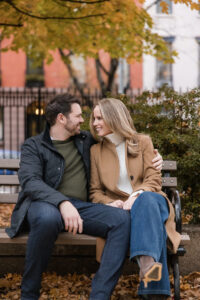A man and a woman sit closely together on a park bench in autumn, smiling at each other during their Chelsea engagement session. Yellow leaves cover the ground and trees behind them.