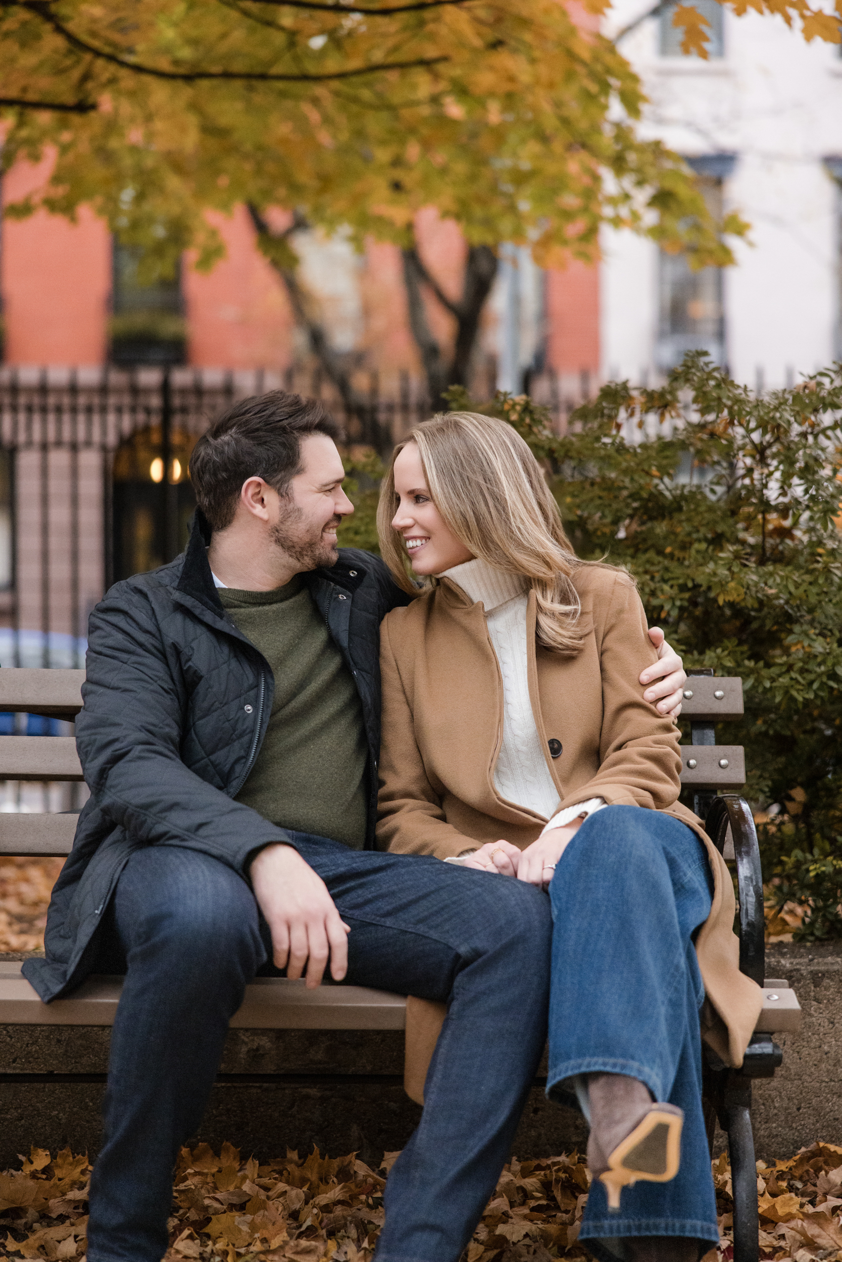 A man and a woman sit closely together on a park bench in autumn, smiling at each other during their Chelsea engagement session. Yellow leaves cover the ground and trees behind them.