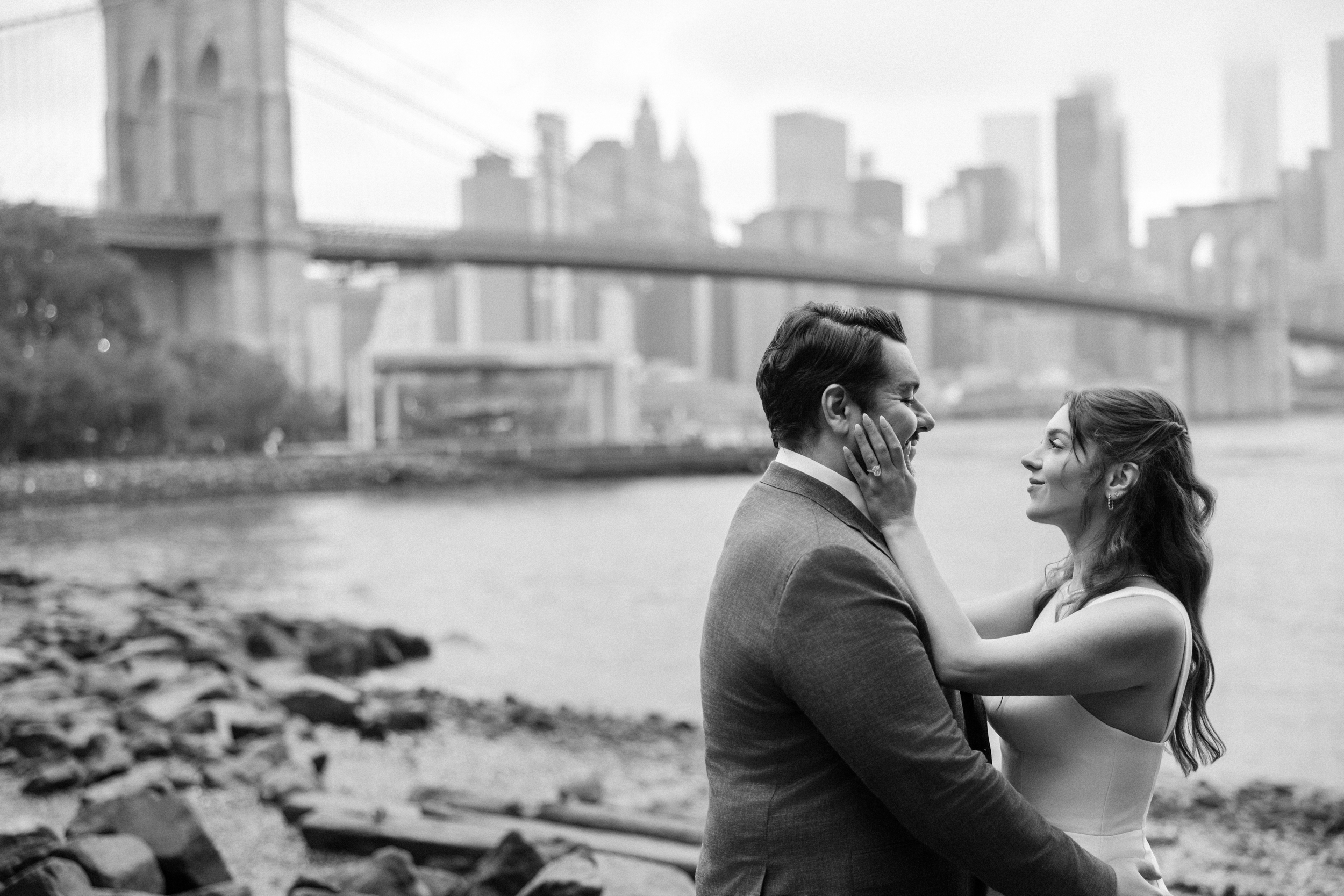 A couple stands close together by the waterfront during a rainy NYC engagement session, with the Brooklyn Bridge and Manhattan skyline in the background on a cloudy day.