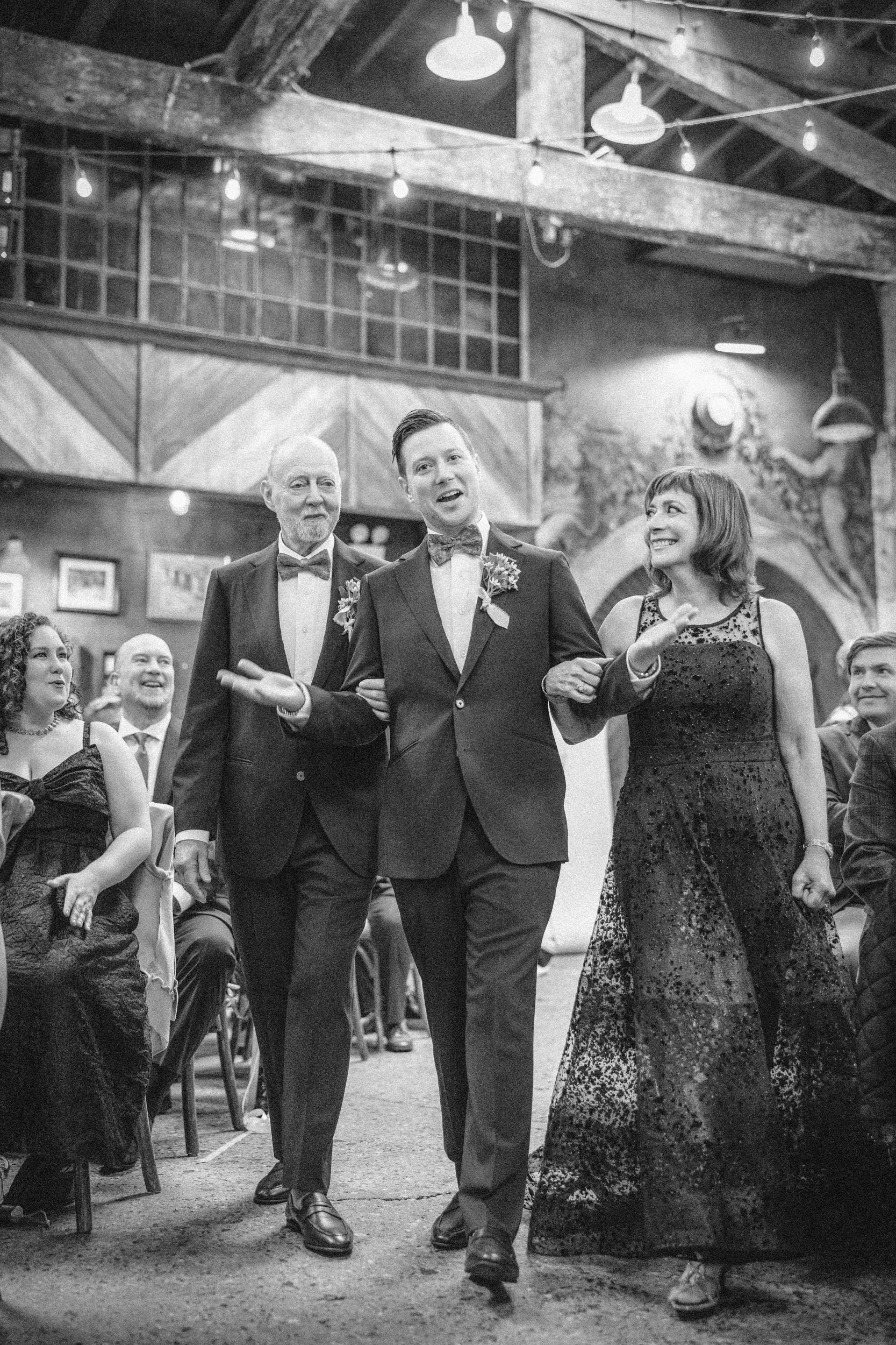 A man in a tuxedo walks arm-in-arm with an older man and woman at a Houston Hall wedding, while guests seated nearby look on.