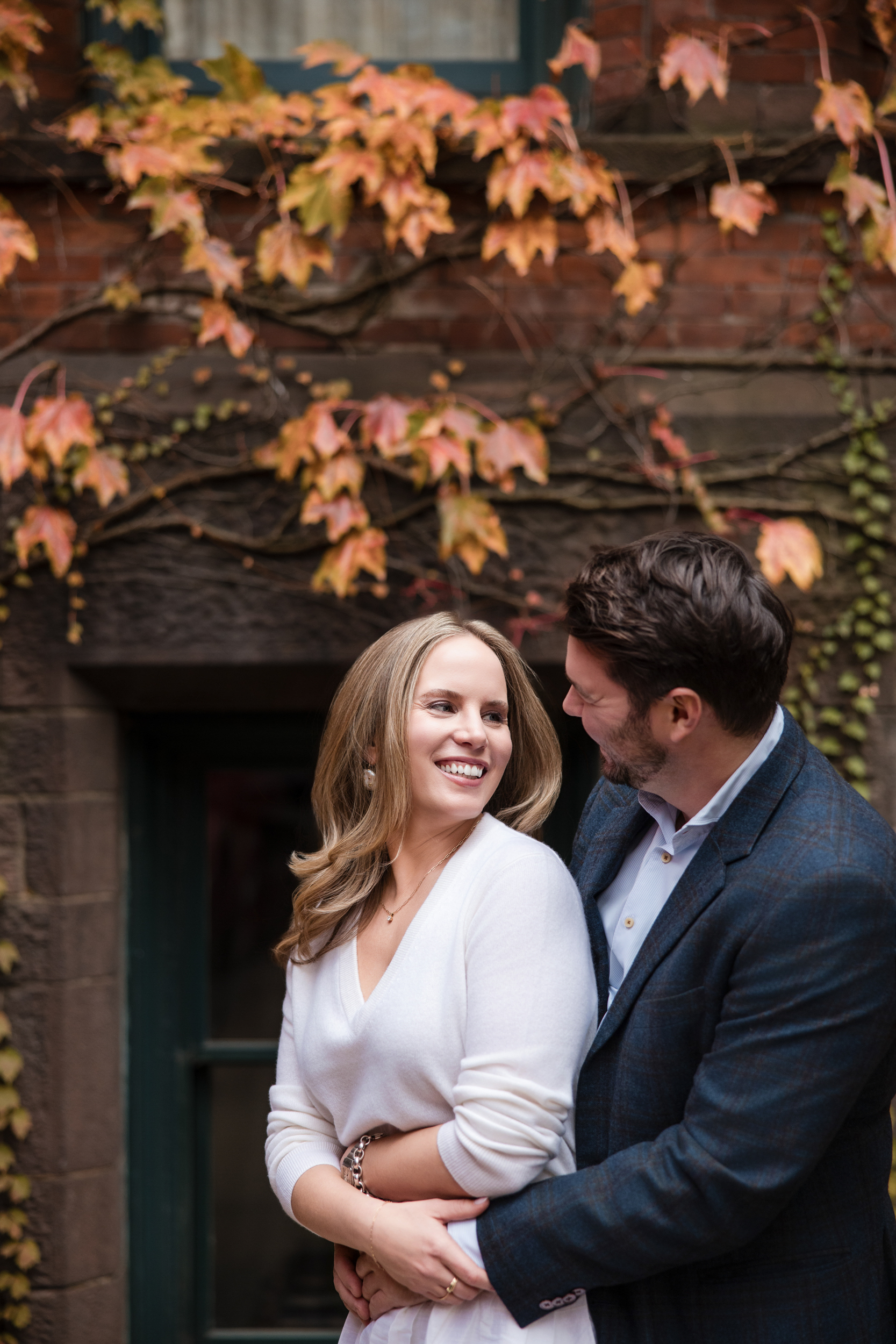 A couple stands close together, smiling at each other in front of a brick wall with autumn leaves and vines during their High Line Hotel Chelsea engagement session.