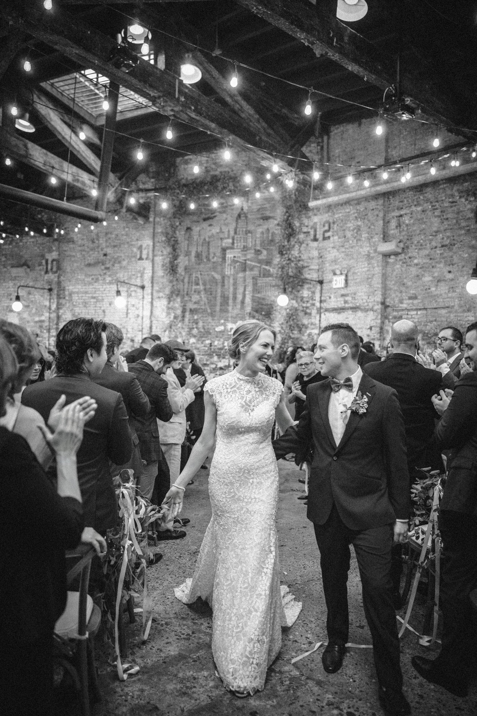 A bride and groom walk down the aisle holding hands, smiling at each other, as guests applaud in a beautifully decorated Houston Hall wedding venue adorned with string lights.
