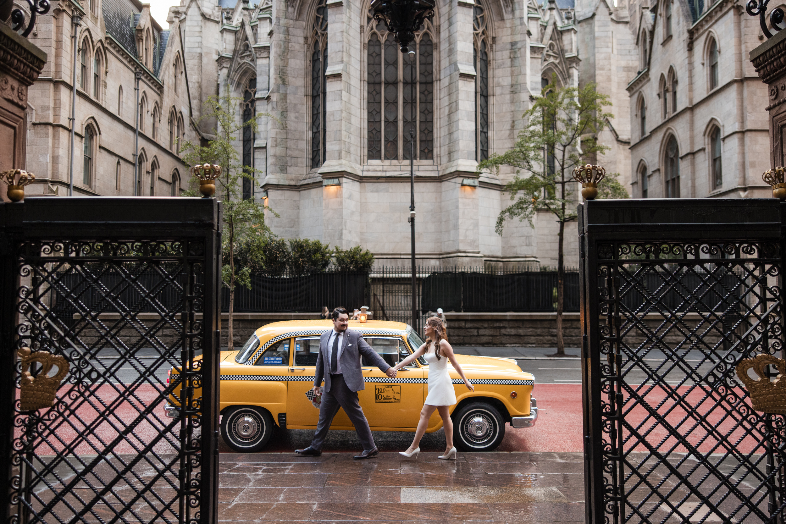 A man in a suit and a woman in a white dress walk past a classic yellow taxi outside a historic stone building, capturing the charm of a rainy NYC engagement session.