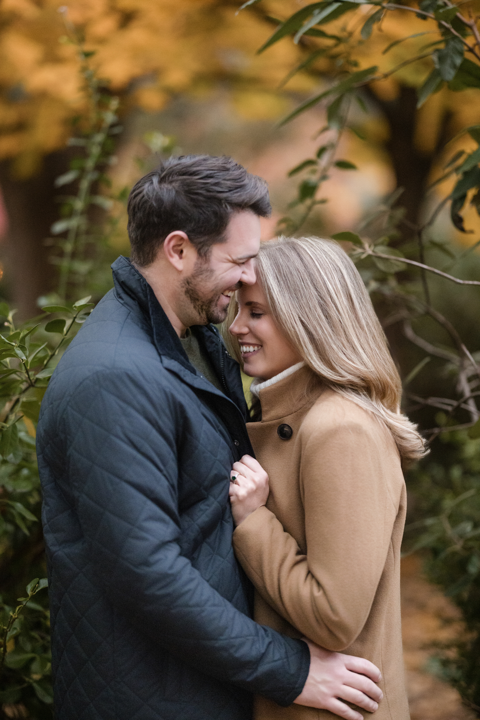 A man and woman stand closely together outdoors during their Chelsea engagement session, smiling and touching foreheads, surrounded by green foliage and autumn-colored trees.