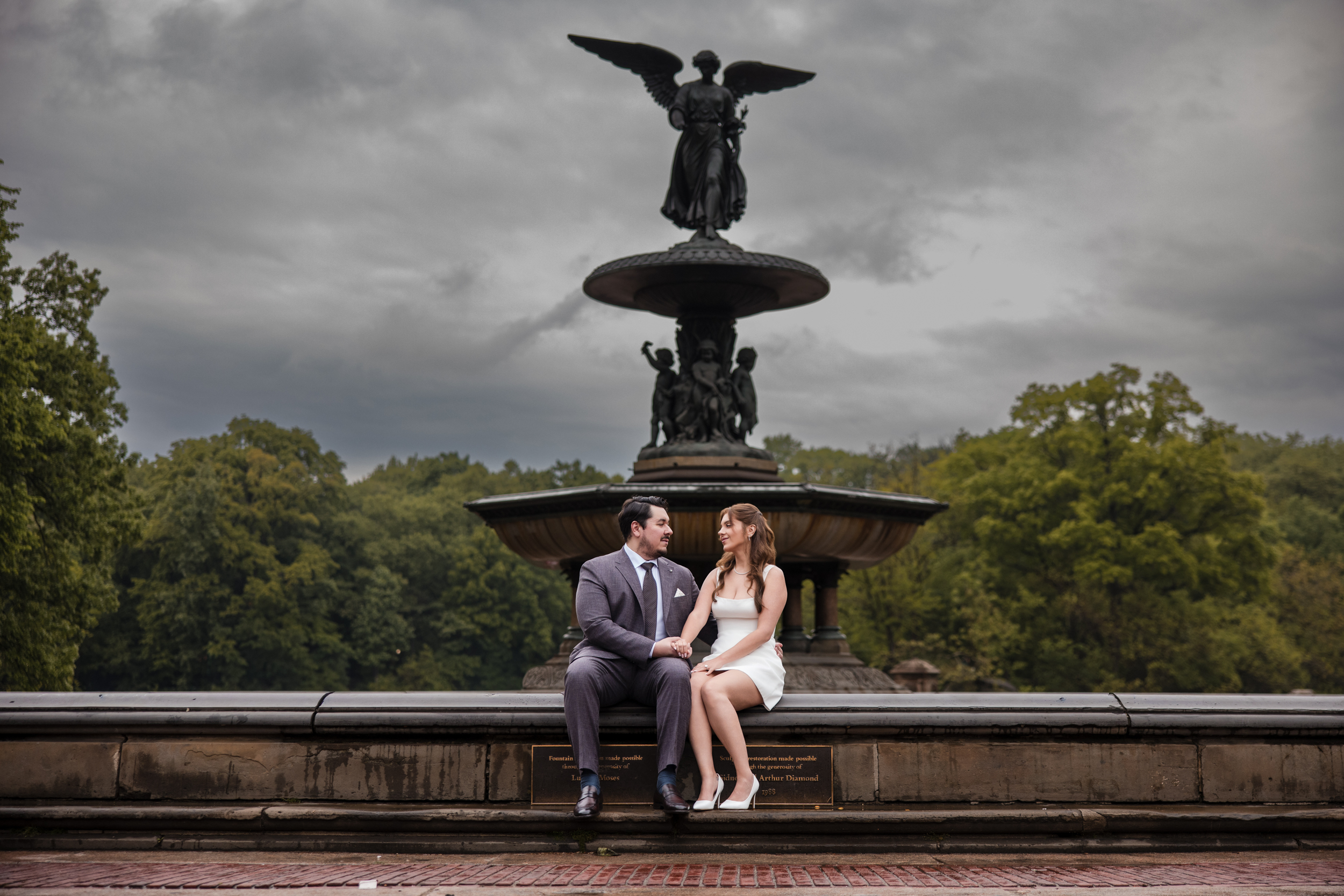 A man in a suit and a woman in a white dress sit together on the edge of Bethesda Fountain in Central Park during their rainy NYC engagement session, with trees and cloudy skies creating a romantic backdrop.