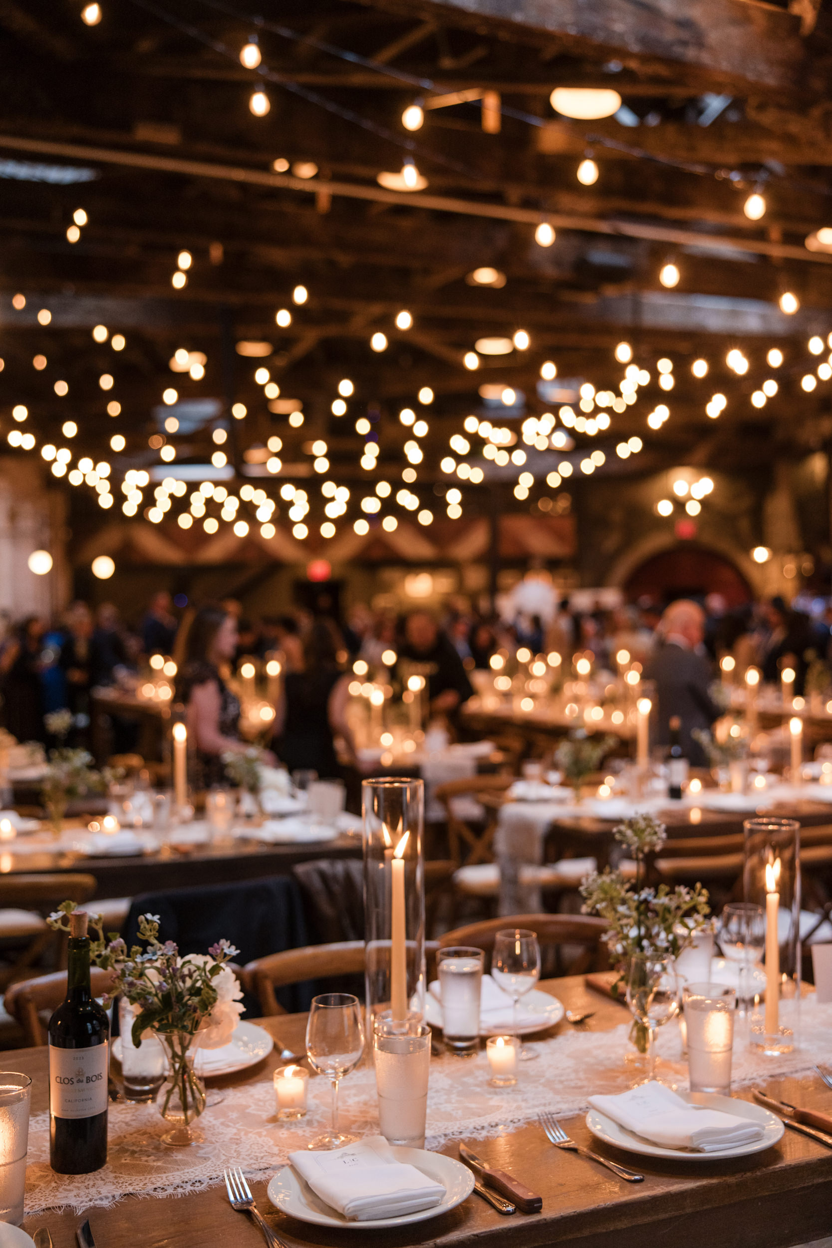 A decorated banquet hall, perfect for a Houston Hall wedding, features string lights overhead, set tables with candles, flowers, wine bottles, and plates—while guests mingle in the background.