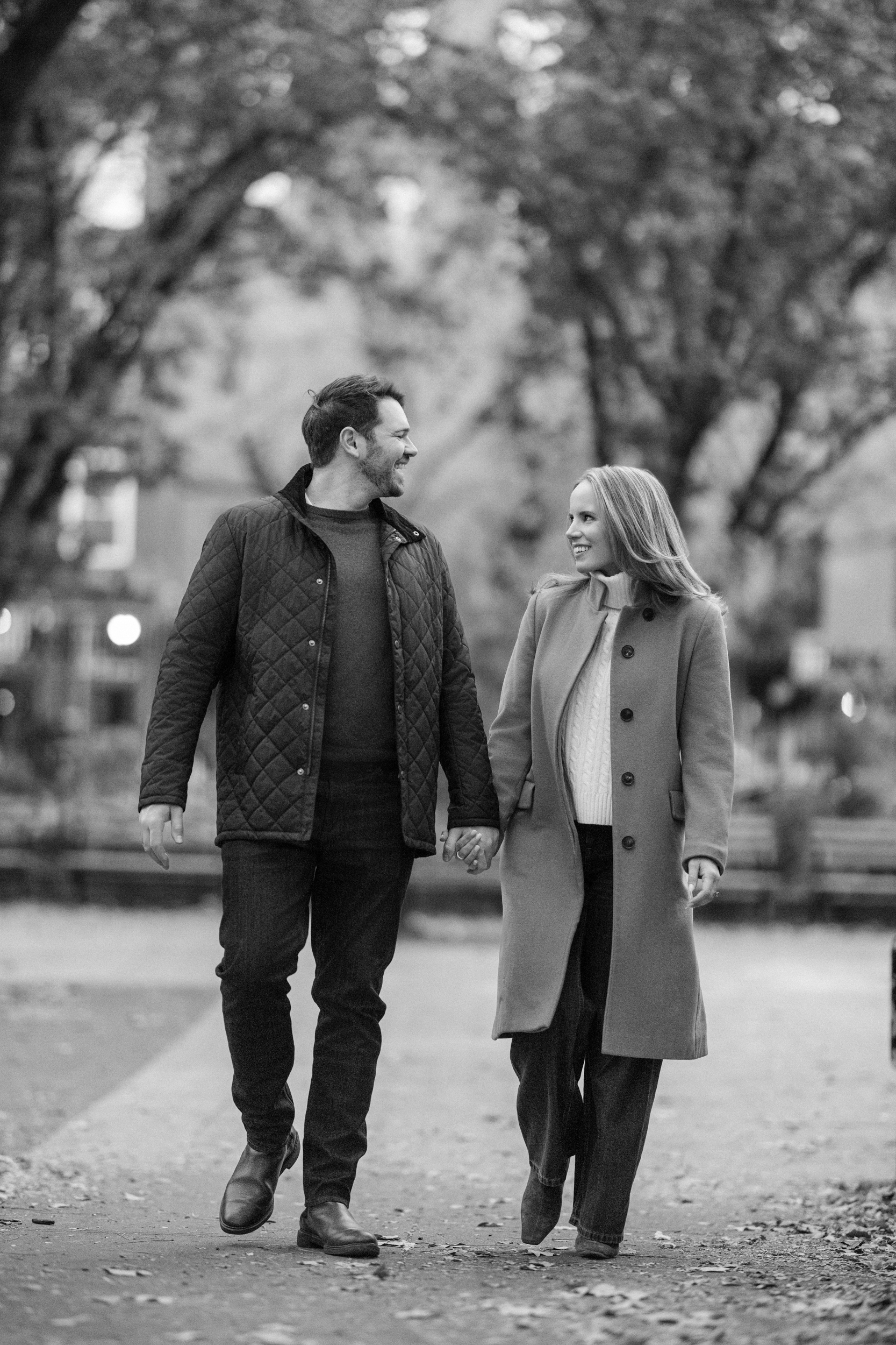 A man and woman walk hand in hand along a tree-lined path, wearing coats and looking at each other during their Chelsea engagement session on an overcast day.
