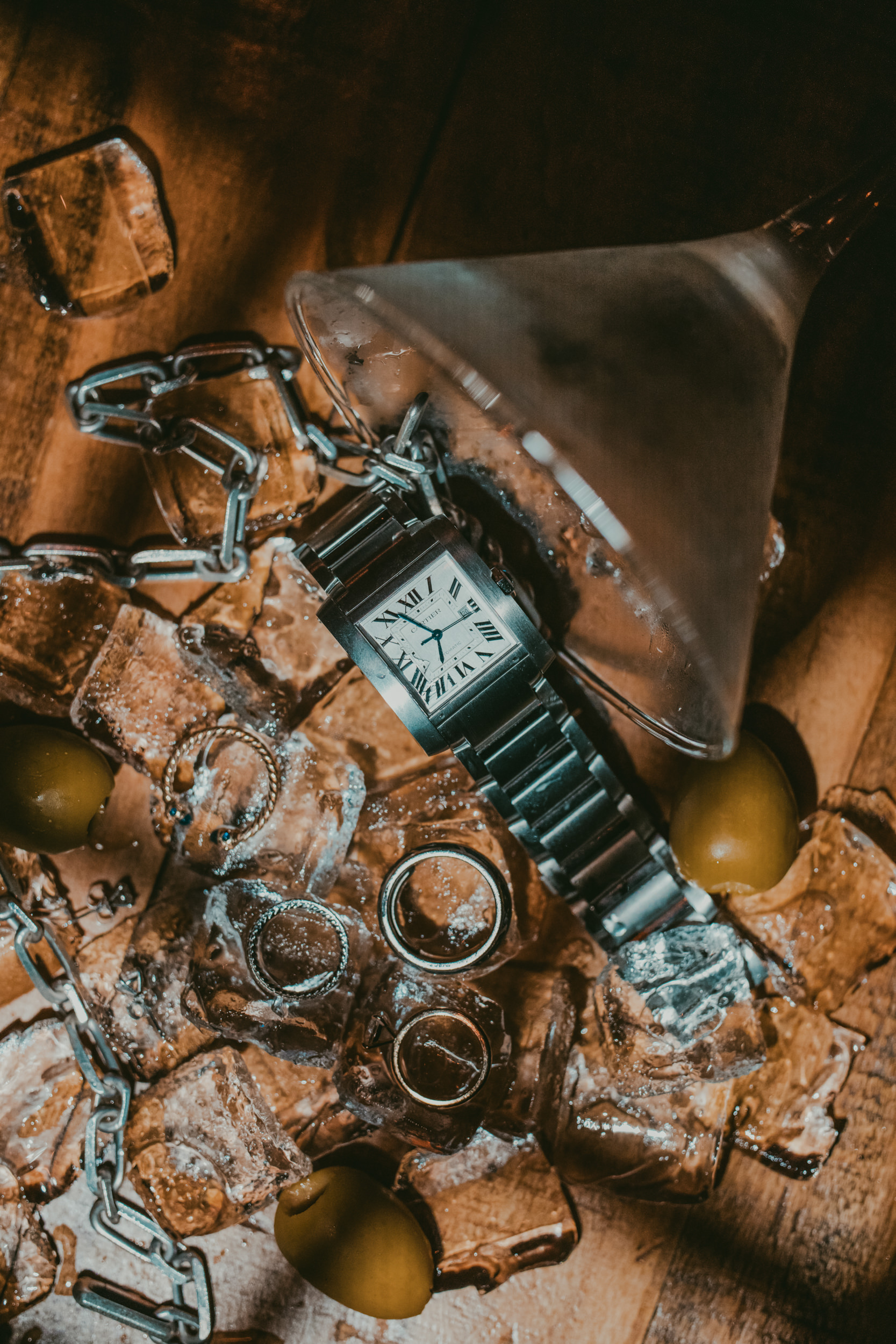 A silver wristwatch, two rings, ice cubes, green olives, and a metal chain are arranged on a wooden surface with a martini glass tipped over—reminiscent of an elegant Houston Hall wedding celebration.