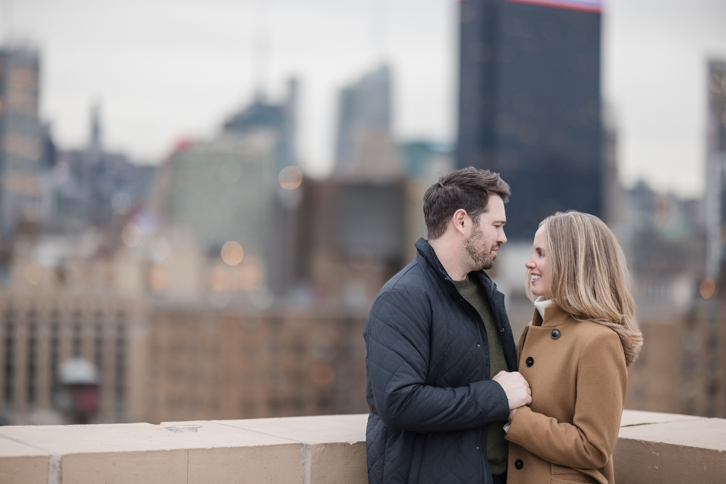 A man and a woman stand close together, holding hands and facing each other during their NYC rooftop engagement session, with a blurred city skyline in the background.