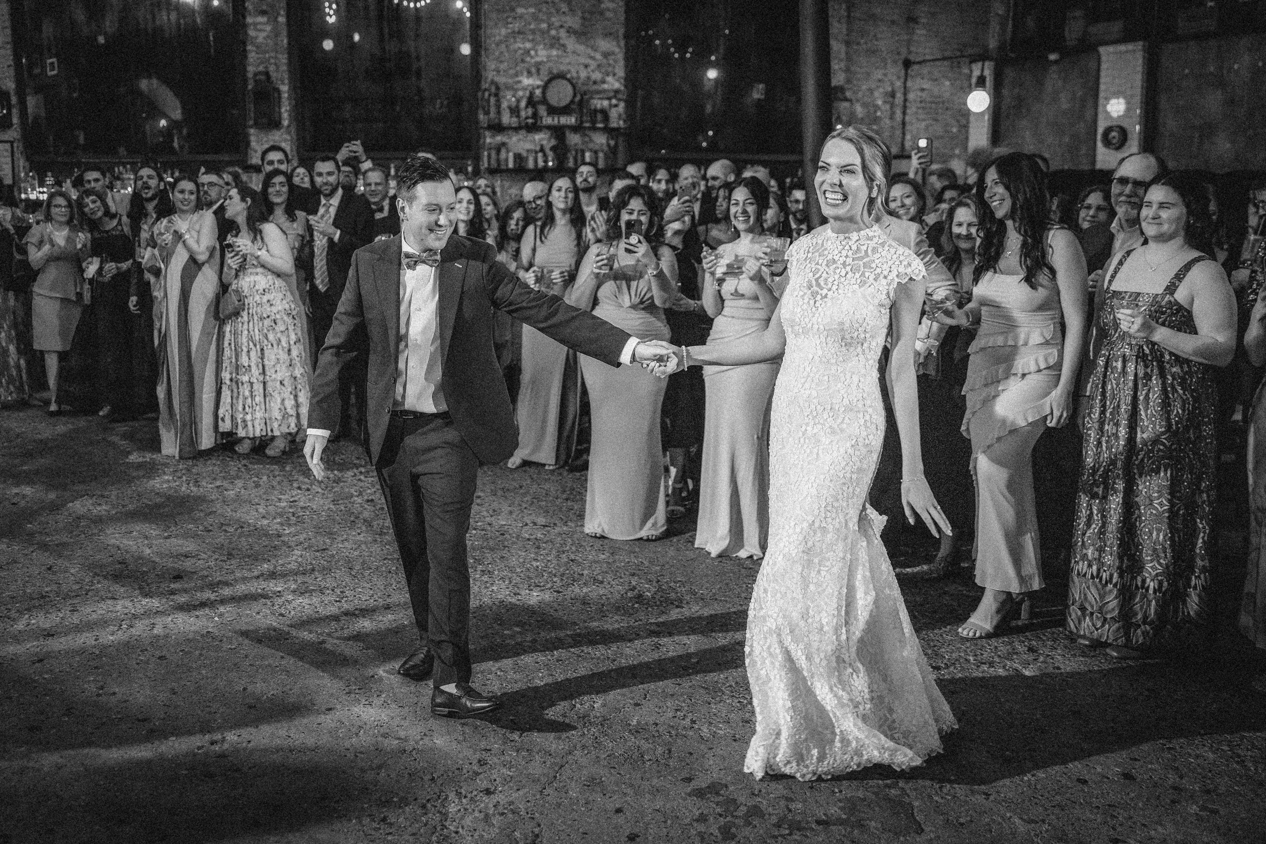 A bride and groom smile as they hold hands and dance, surrounded by guests watching and taking photos at a beautiful Houston Hall wedding in a large indoor venue.