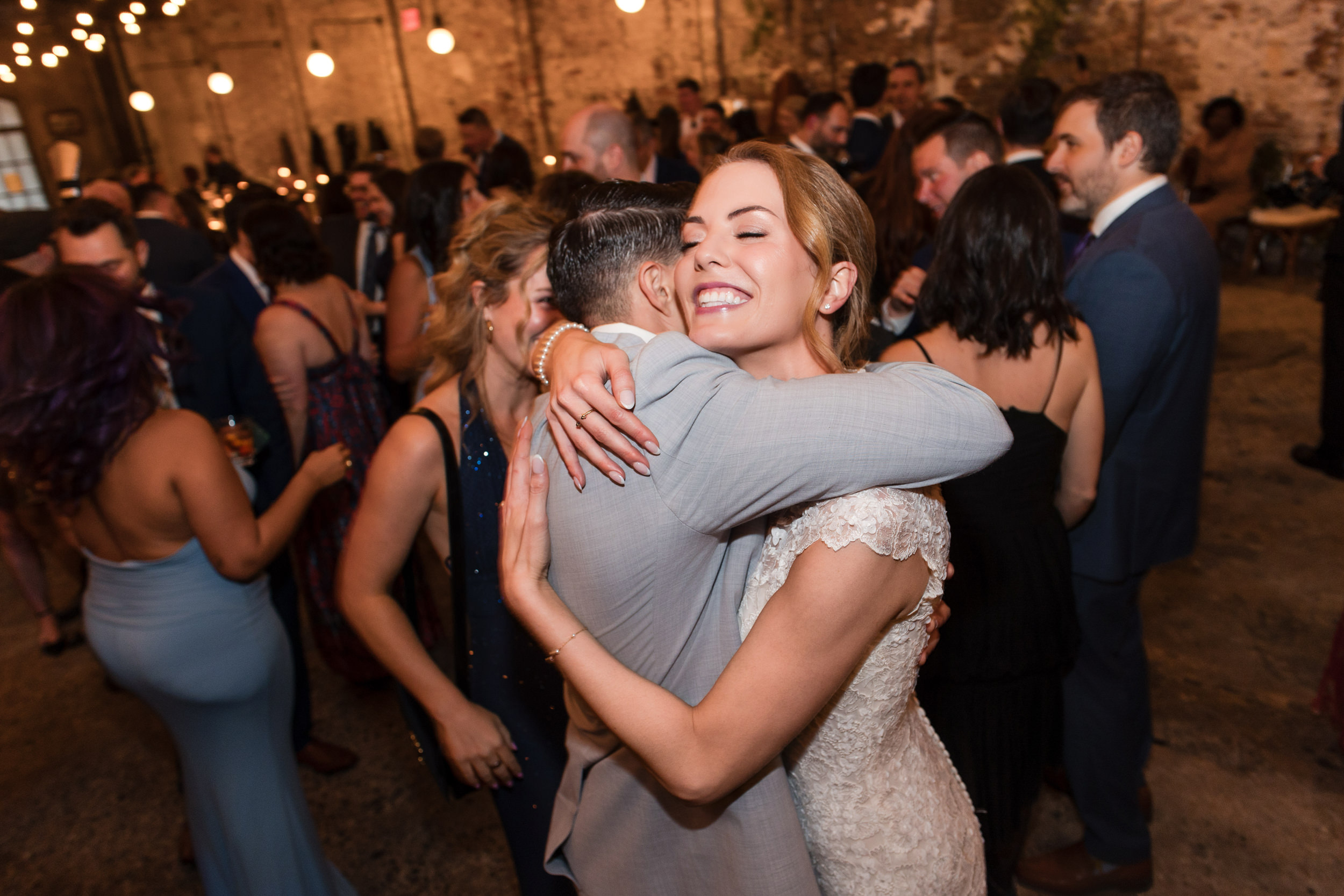 A bride in a white dress hugs a man in a suit while smiling, surrounded by guests at a Houston Hall wedding reception with string lights and exposed brick walls.