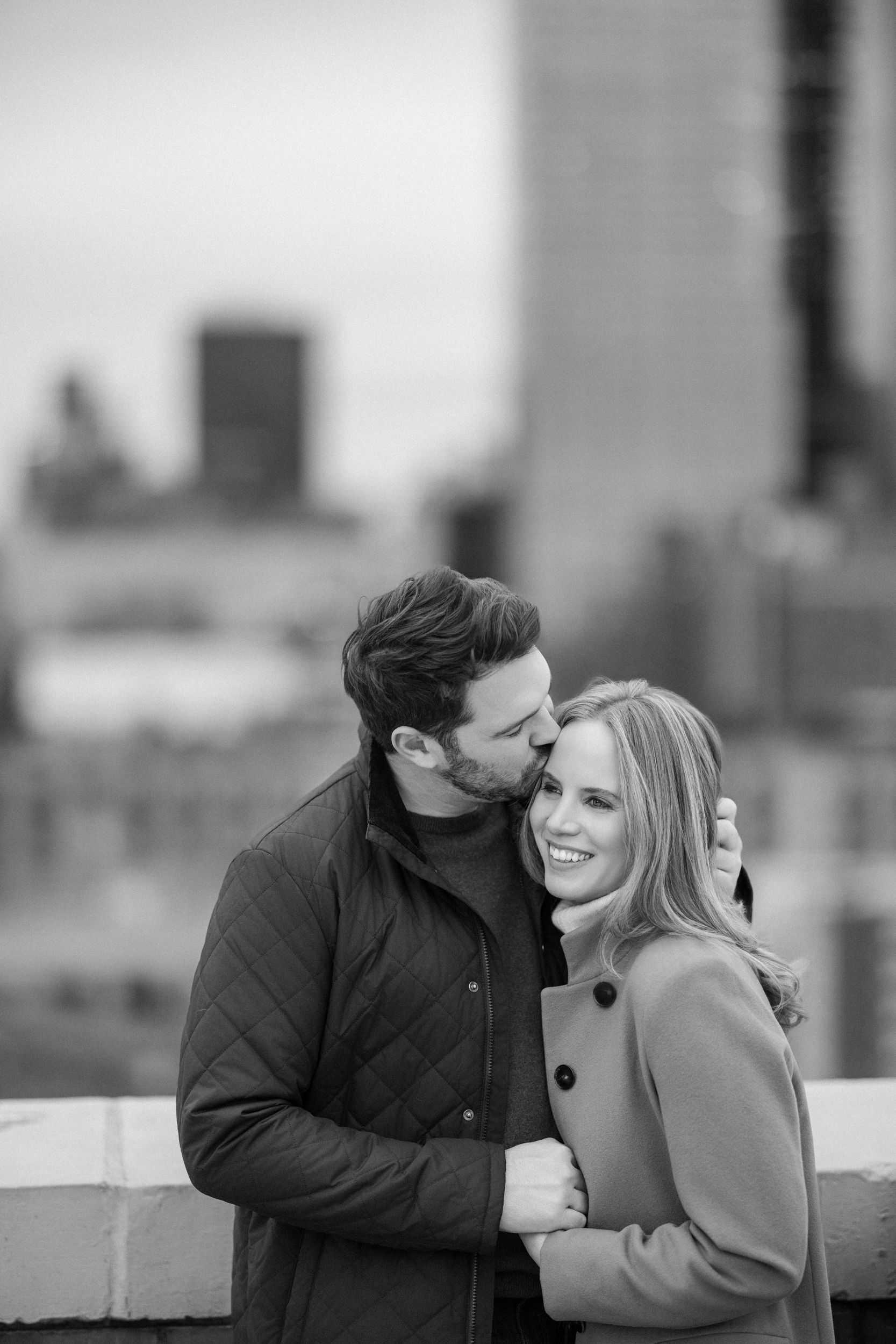 A man kisses a smiling woman on the forehead while standing together outdoors during an NYC rooftop engagement session, with a blurred cityscape in the background.
