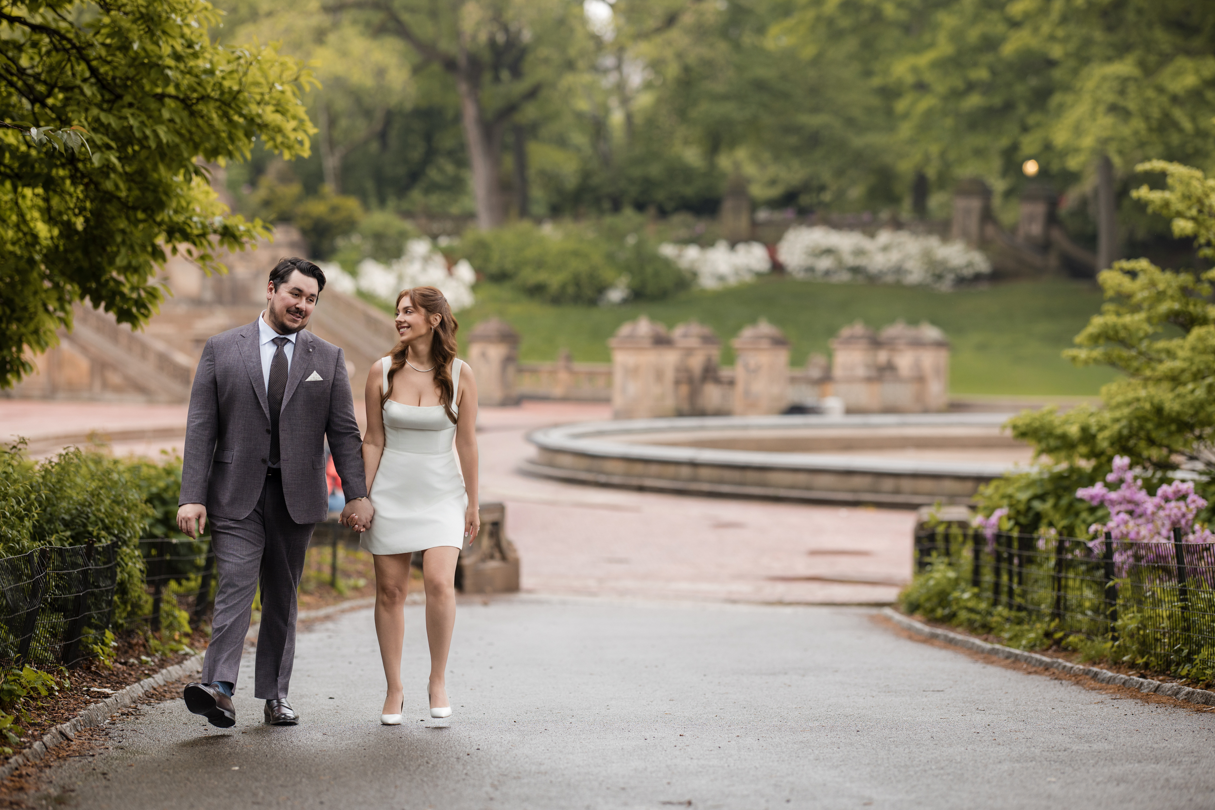 A man in a suit and a woman in a white dress walk hand in hand on a paved path in a park, smiling at each other during their rainy NYC engagement session, with lush greenery and a fountain in the background.