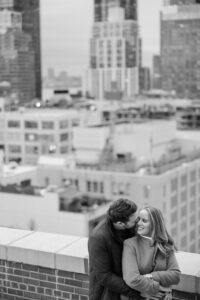 A man and woman stand close together on a NYC rooftop during their engagement session, with tall buildings in the background. The man leans toward the woman's cheek while she smiles. The image is in black and white.