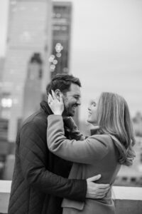 A couple stands close together on a NYC rooftop during their engagement session, smiling at each other with city buildings blurred in the background.