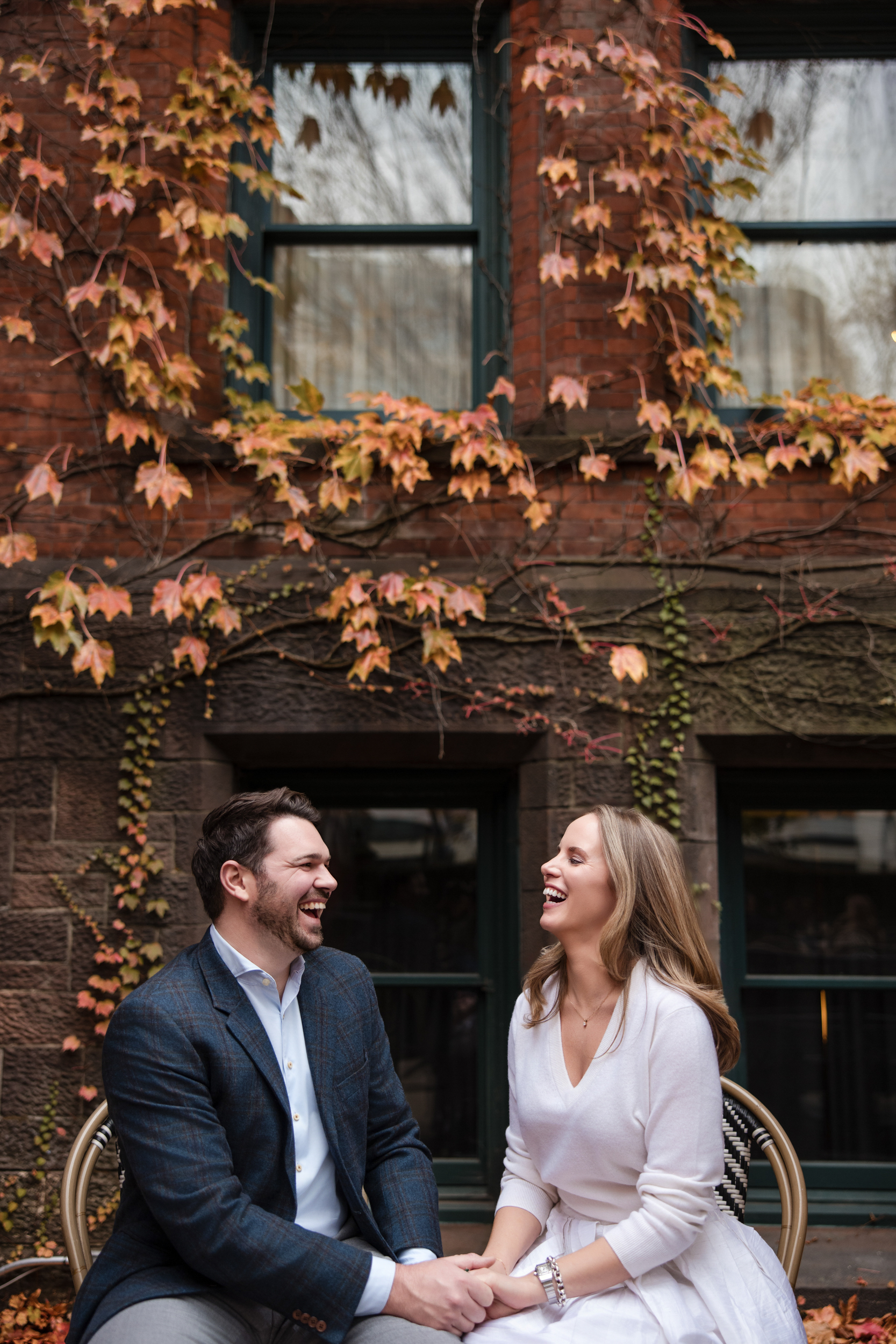 A man and woman sit outdoors in front of a brick building with ivy, holding hands and laughing together during their Chelsea engagement session.