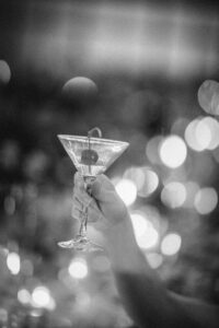 A hand holds up a martini glass with a cocktail and cherry garnish, set against the bokeh-lit backdrop of a festive Houston Hall wedding.