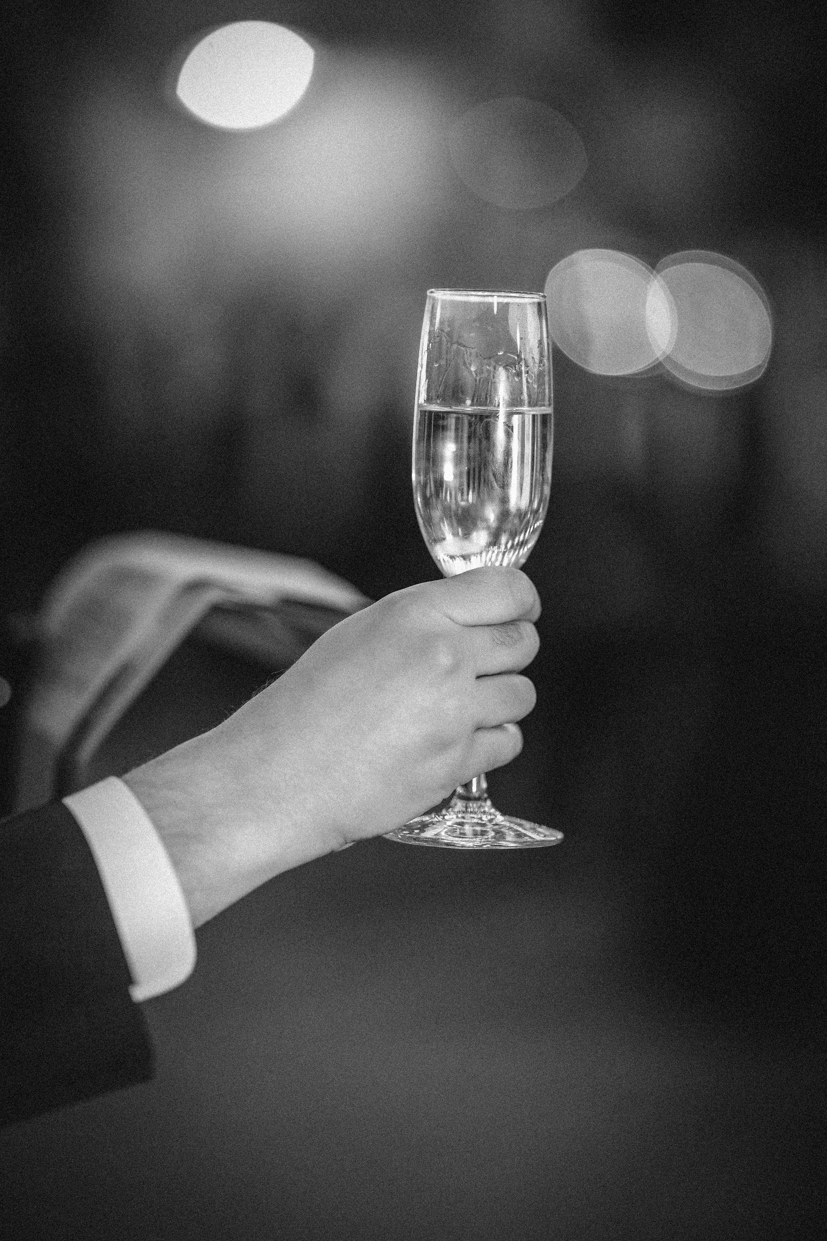 A person in formal attire holds up a champagne flute filled with a clear liquid, celebrating amid the glowing lights of a Houston Hall wedding.