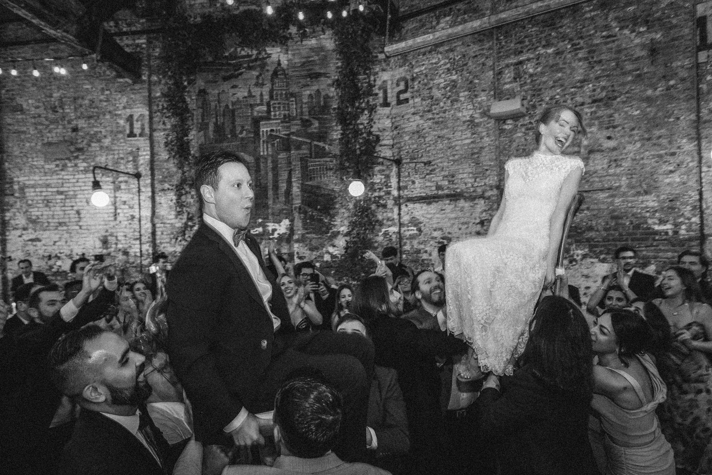A bride and groom are lifted on chairs by guests during a joyful Houston Hall wedding celebration in a large, rustic venue with exposed brick walls.