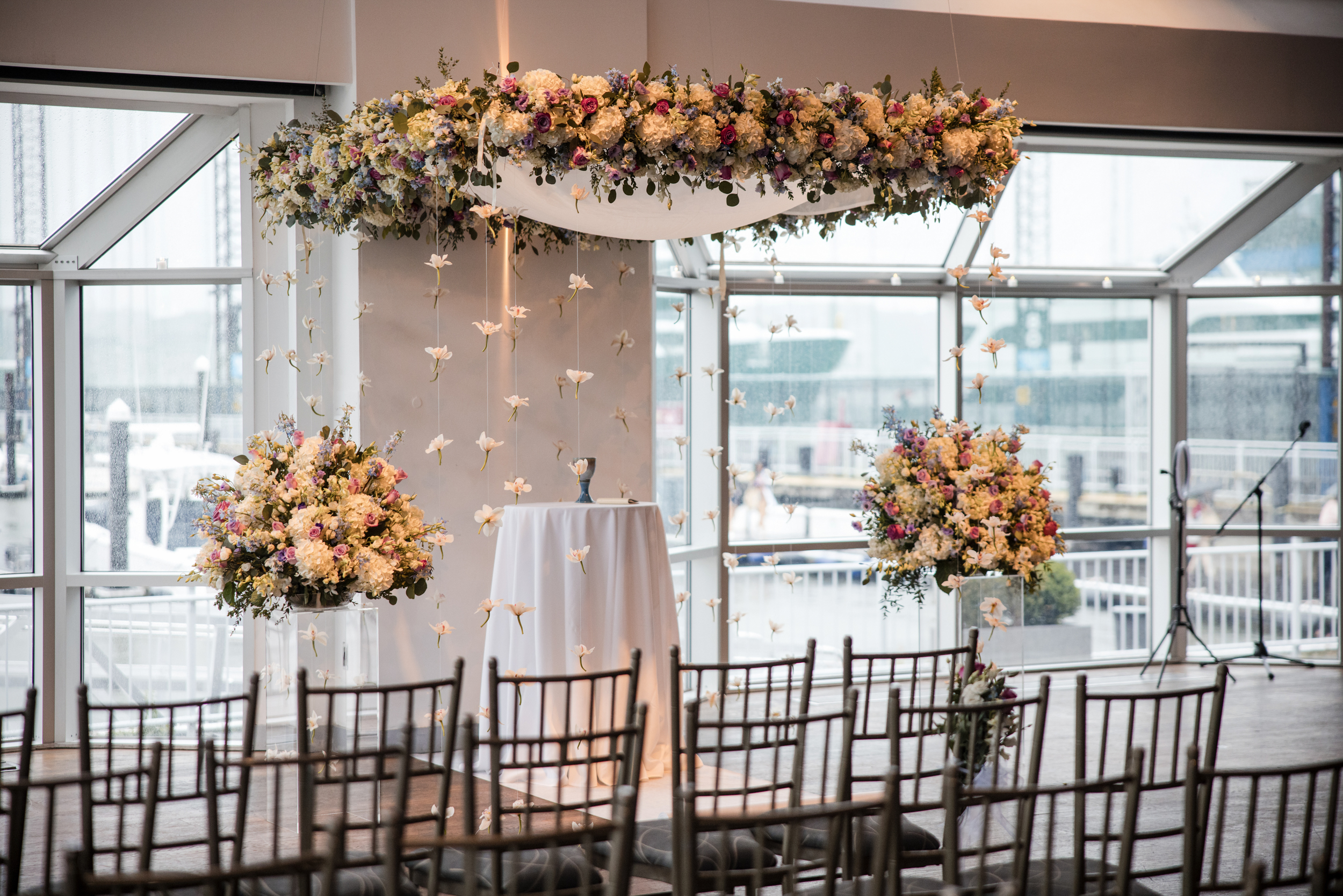 Indoor Pier Sixty and Current Wedding ceremony setup with rows of chairs, a floral arch with hanging flowers, and elegant floral arrangements near a front table, all in a venue filled with light from large windows.