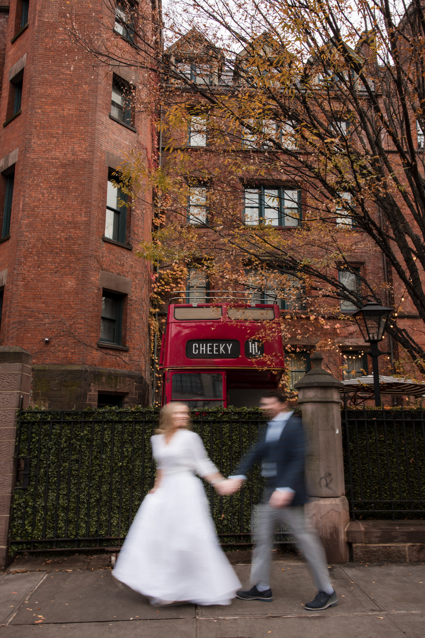 A bride and groom holding hands walk past a red double-decker bus with "CHEEKY" on it, near autumn trees and a brick building during their High Line Hotel Chelsea engagement session.