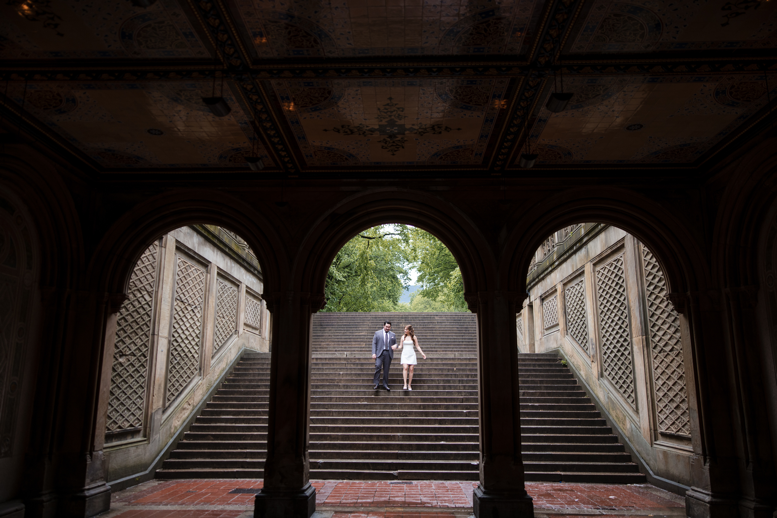 A couple stands together on the middle steps of an outdoor staircase, framed by three large arches and decorative ceilings during their rainy NYC engagement session.