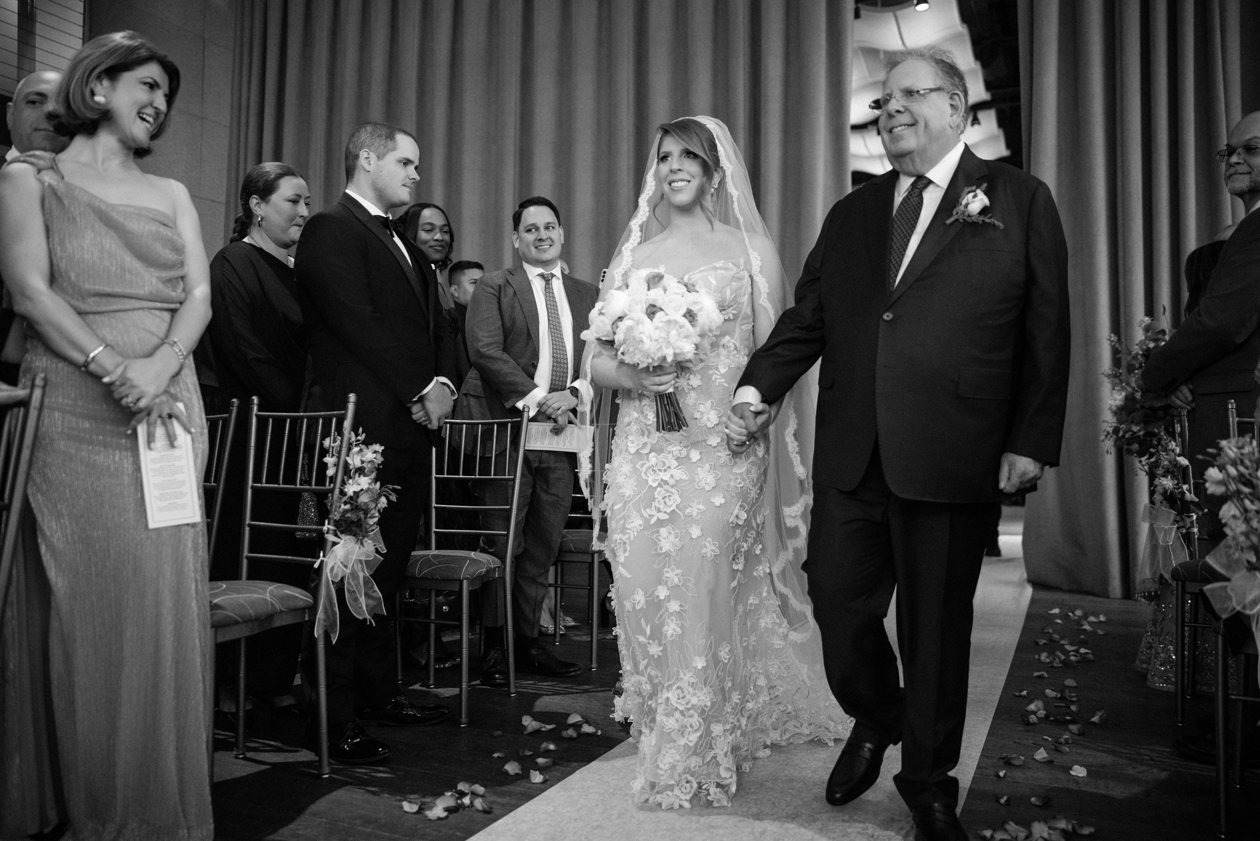 A bride in a lace wedding dress walks down the aisle at her Pier Sixty and Current Wedding, holding hands with an older man, surrounded by smiling guests seated on either side.