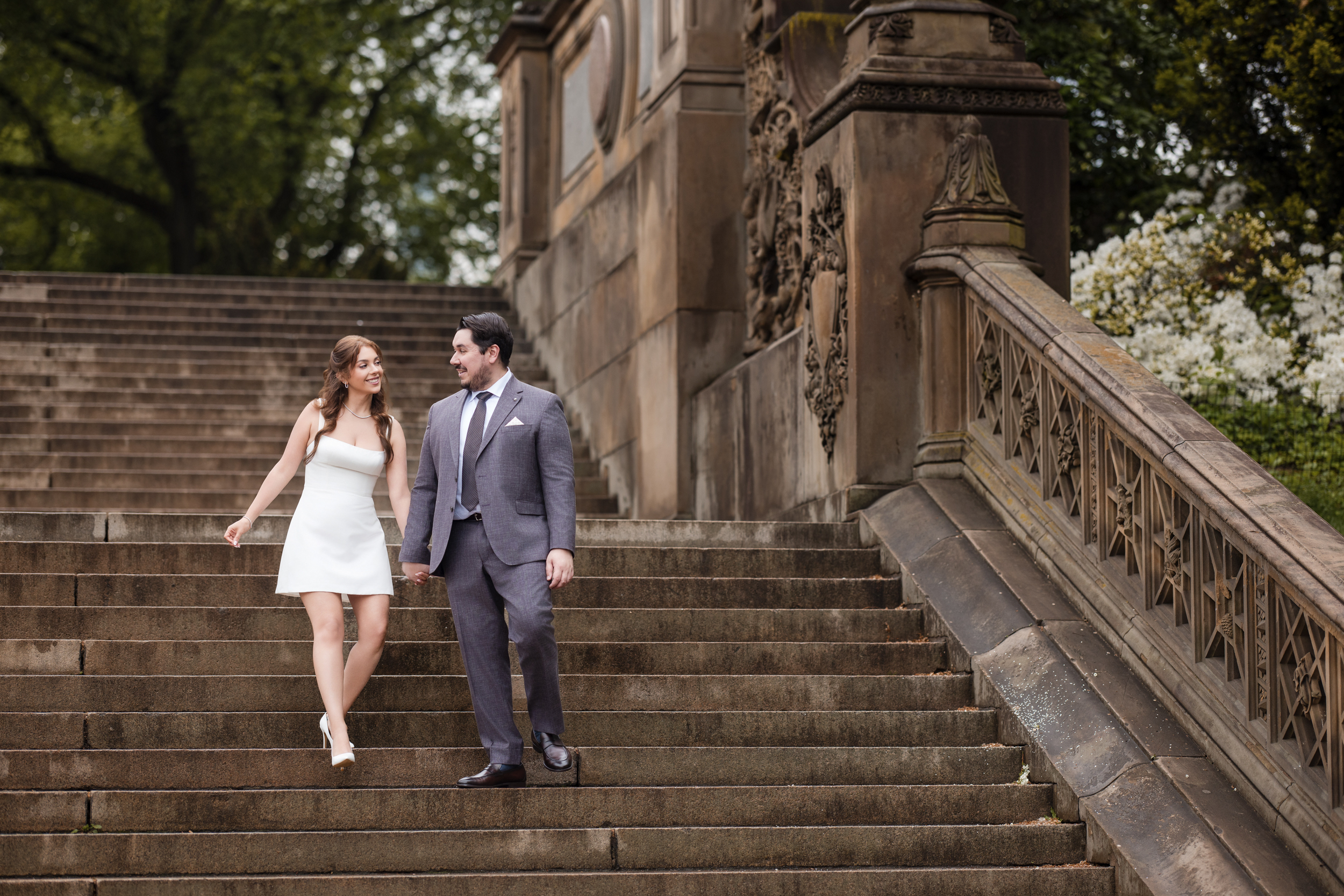 A woman in a white dress and a man in a gray suit walk down stone steps outdoors, holding hands near an ornate structure surrounded by greenery during their rainy NYC engagement session.