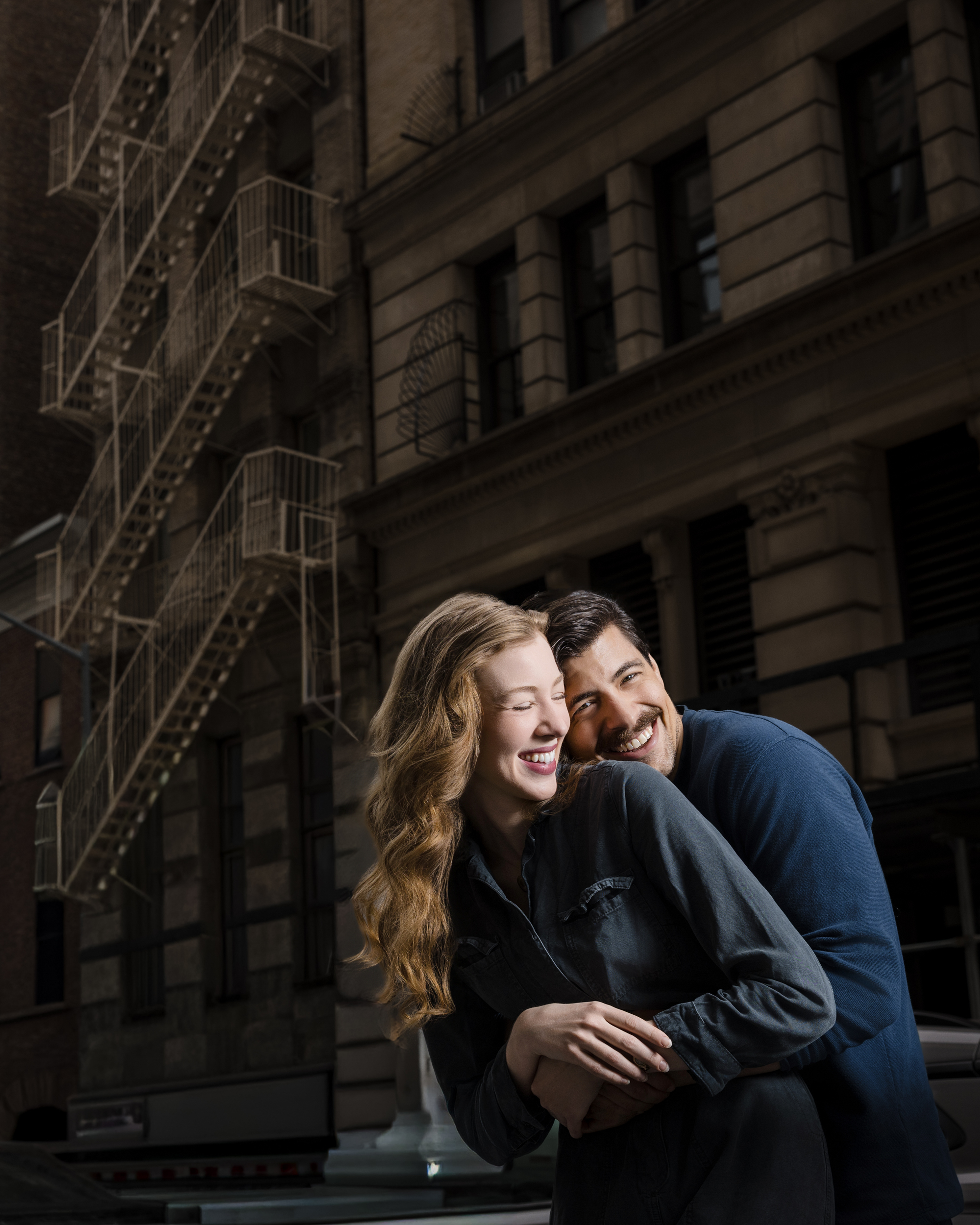 A man and woman stand close together, smiling and laughing, in front of a city building with a metal fire escape during an NYC photo walk.